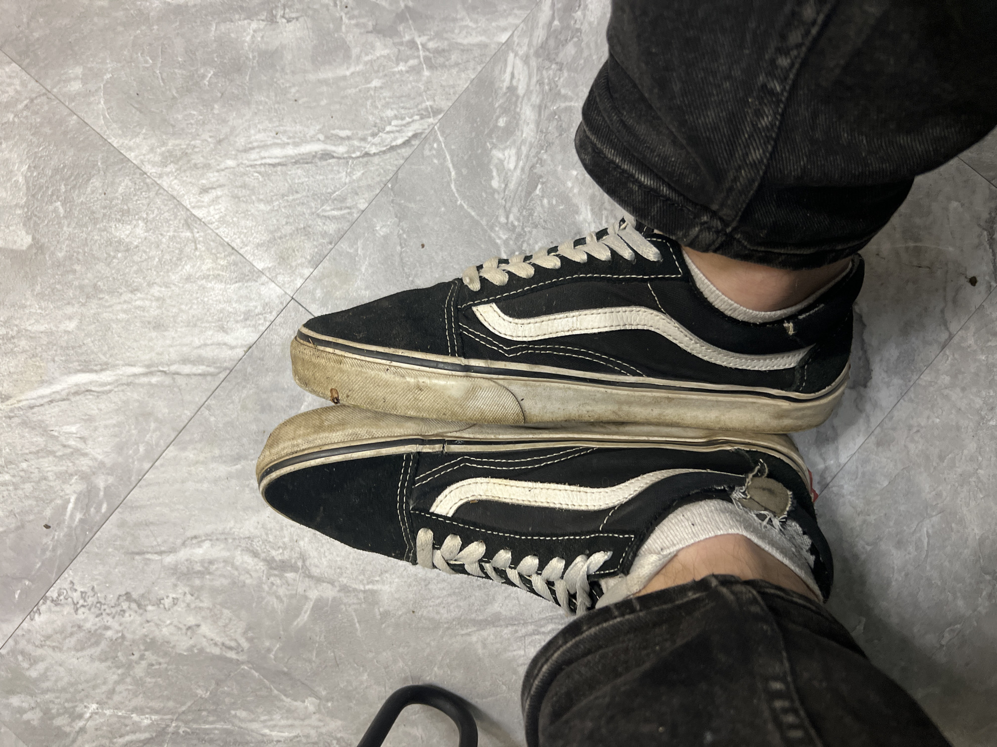 The image displays a pair of black and white Vans Old Skool sneakers worn by an individual whose lower legs and feet are visible. The shoes are positioned side-by-side, with the laces tied. The right shoe shows more wear and dirt on its white sole and side stripe compared to the left shoe. The individual is wearing dark denim jeans that partially cover the shoes, and white socks, one of which appears ripped. The shoes are resting on a light gray tiled floor with visible grout lines. The overall scene suggests a casual indoor environment, possibly a home or a public space with similar flooring. There is no information about the location in the city of Ленинский, Russia that can be inferred from the image.