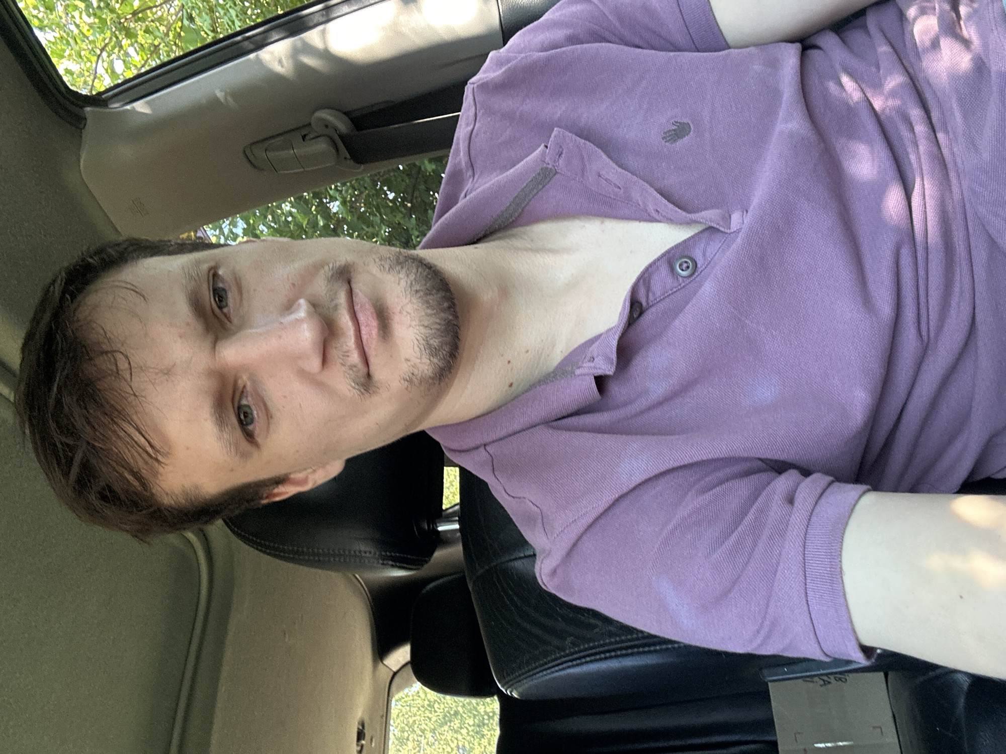 A man is sitting in the driver's seat of a car, looking at the camera. He has short brown hair and a light beard. He is wearing a purple polo shirt. The car interior is visible behind him, with black leather seats. Outside the car, green trees and foliage are visible. The image appears to have been taken during the daytime, with sunlight filtering through the trees and casting shadows on the man and the car's interior. There is a cardboard box on the passenger seat behind him. The image does not contain information that would allow for an accurate determination of the location within the city of Shatava, Ukraine, or any other specific city.