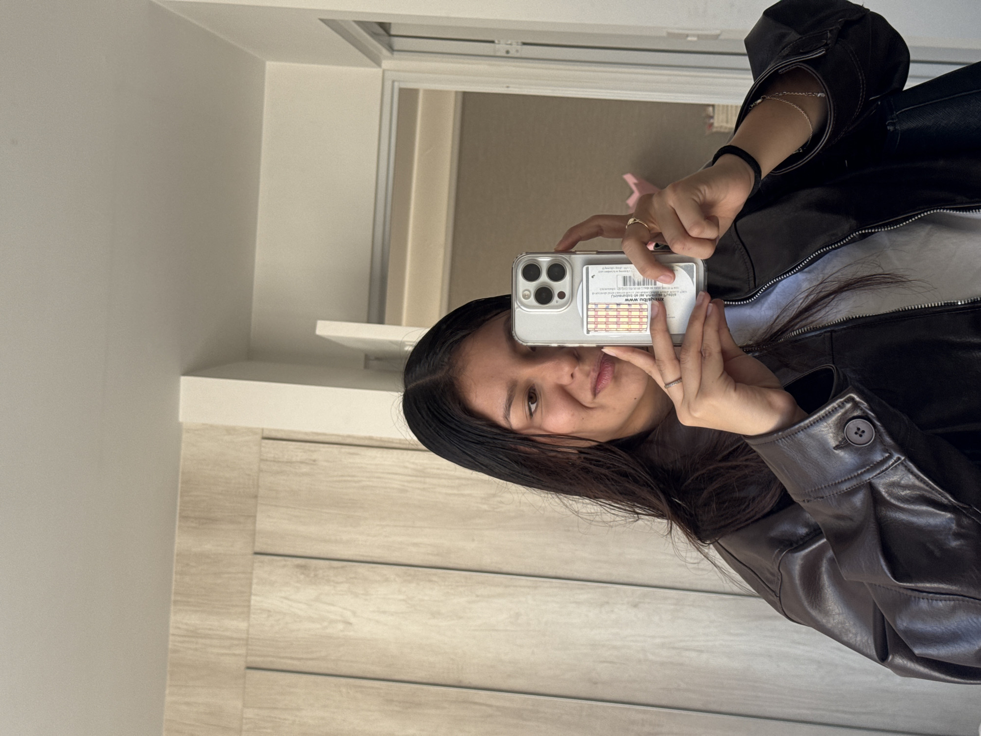 A young woman is depicted taking a mirror selfie with her smartphone, captured from an upward angle. She has long, dark hair and a subtle smile. She is wearing a black leather jacket over a light-colored garment. Accessories include a ring on her right ring finger, a thin silver bracelet on her left wrist, and a black hair tie also on her left wrist.
She holds a silver smartphone, likely an iPhone given the prominent camera module, in a clear protective case. On the back of the phone case, there is a white sticker or card with a barcode and text. Clearly visible in the mirror's reflection, the text reads "Ubuy www.ubuy.com.mx".
The setting appears to be an indoor room with light-colored, possibly off-white walls. To the left, there is light-toned wooden paneling. The mirror itself has a thin white frame. In the reflection behind the woman, part of a doorway or opening is visible, along with a blurred pink object. The room is brightly lit, suggesting either natural daylight or well-distributed artificial light. The activity is a casual mirror selfie. The location context for this media is San Francisco Ocotlán (Ocotlán), Mexico.