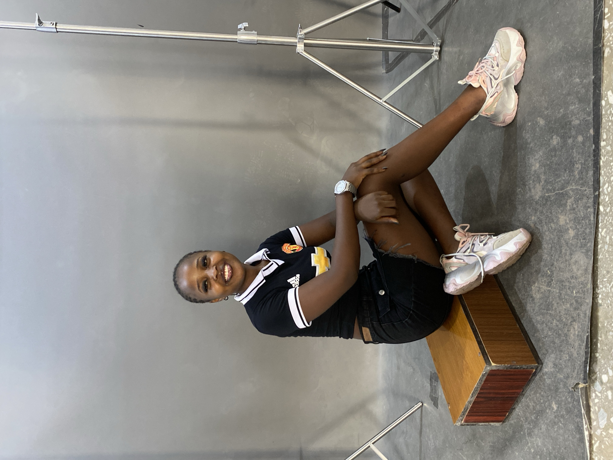 A young Black woman with braided hair and full makeup sits on a wooden box in front of a gray backdrop. She wears a black Adidas t-shirt with white trim on the collar and sleeves, and a black denim skirt with frayed edges. She has a silver watch on her left wrist and light-colored sneakers with pink accents. The woman is smiling and looking towards the camera. The background is a plain gray studio backdrop with a lighting stand visible on the right side of the image. The floor appears to be concrete. There are no indications of the location being in Gura Top, Nigeria.