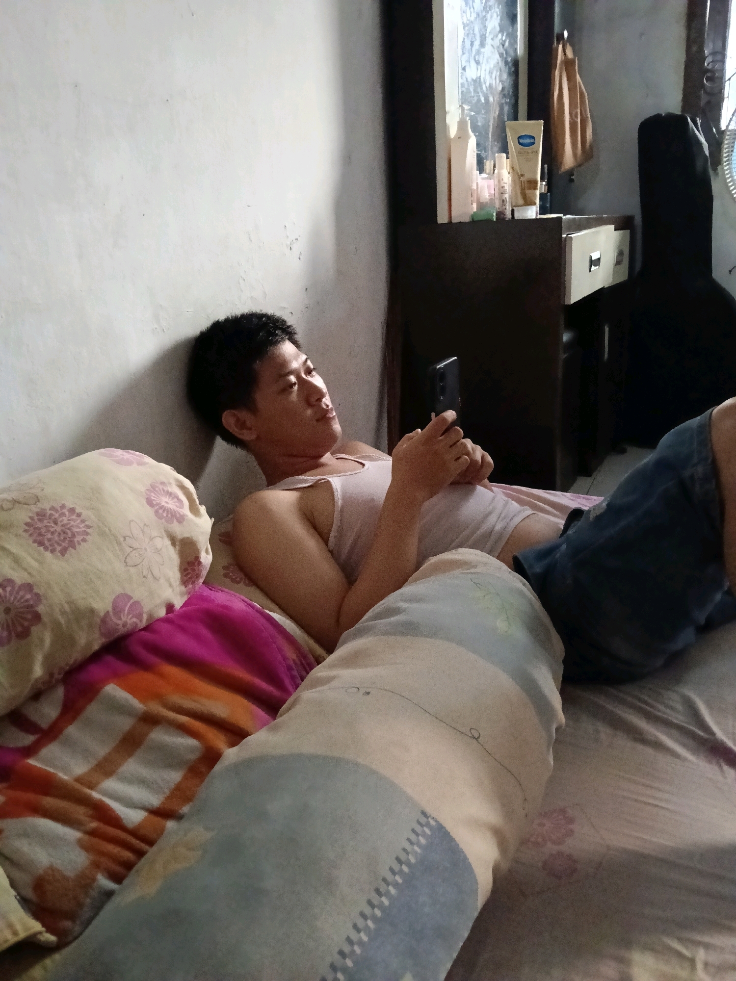 The image depicts a young Asian male lying on a bed in a room. He is wearing a light-colored tank top and blue shorts. The man is holding a smartphone in his right hand, appearing to be engaged with it. He is positioned near a wall, and several pillows are visible on the bed around him. In the background, a dresser with various items on top, including bottles and a bag, is present. A black guitar case is also visible to the right of the dresser. The room appears to be an indoor residential space.