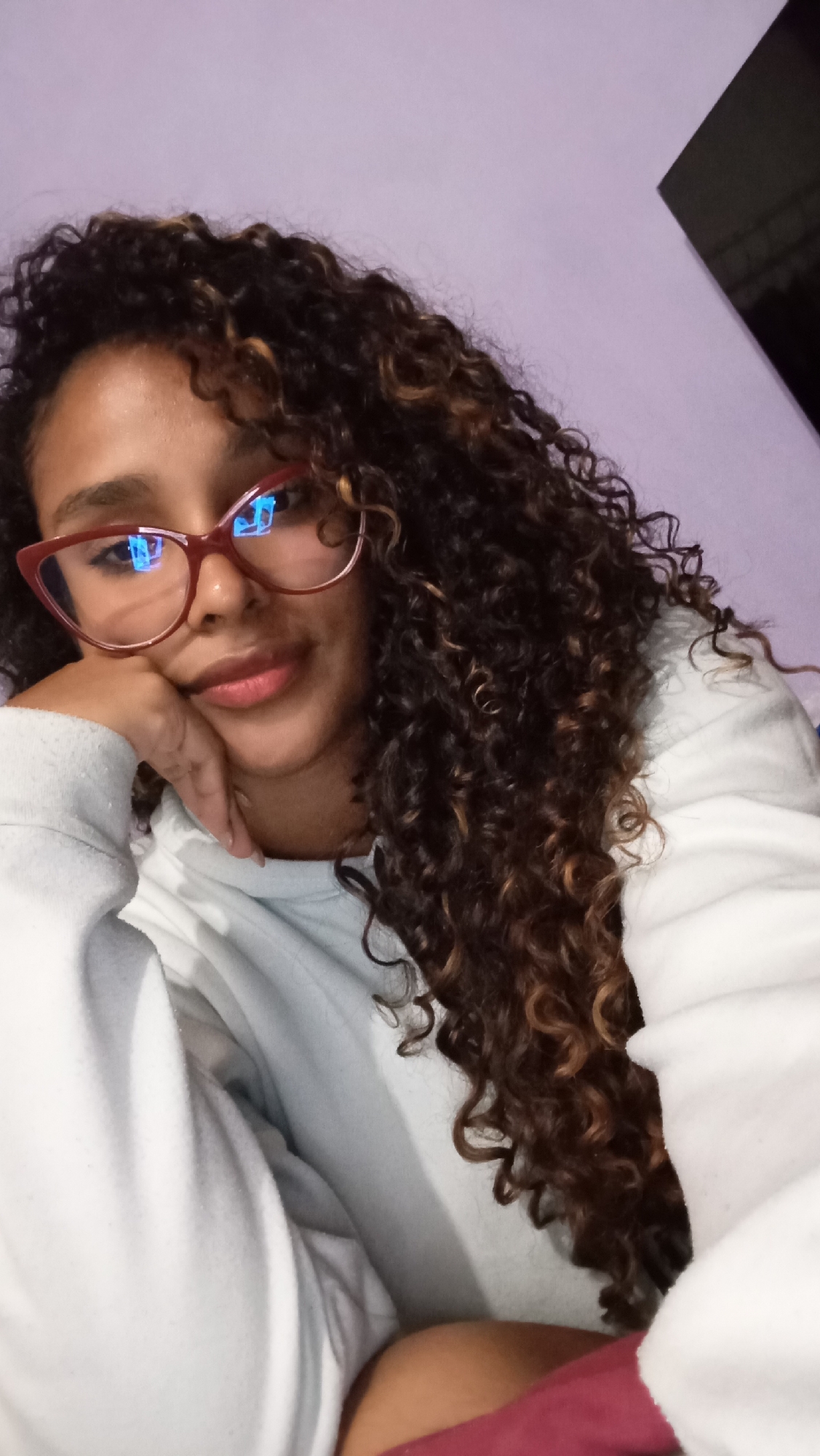 A woman with curly brown and dark brown hair and wearing red glasses is looking at the camera, her head resting on her hand. She is wearing a light-colored hoodie. The background is a plain light purple wall. A dark object is visible in the upper right corner of the frame. There is no specific information to suggest this image was taken in Juan Griego, Venezuela. The location can not be verified in the image.