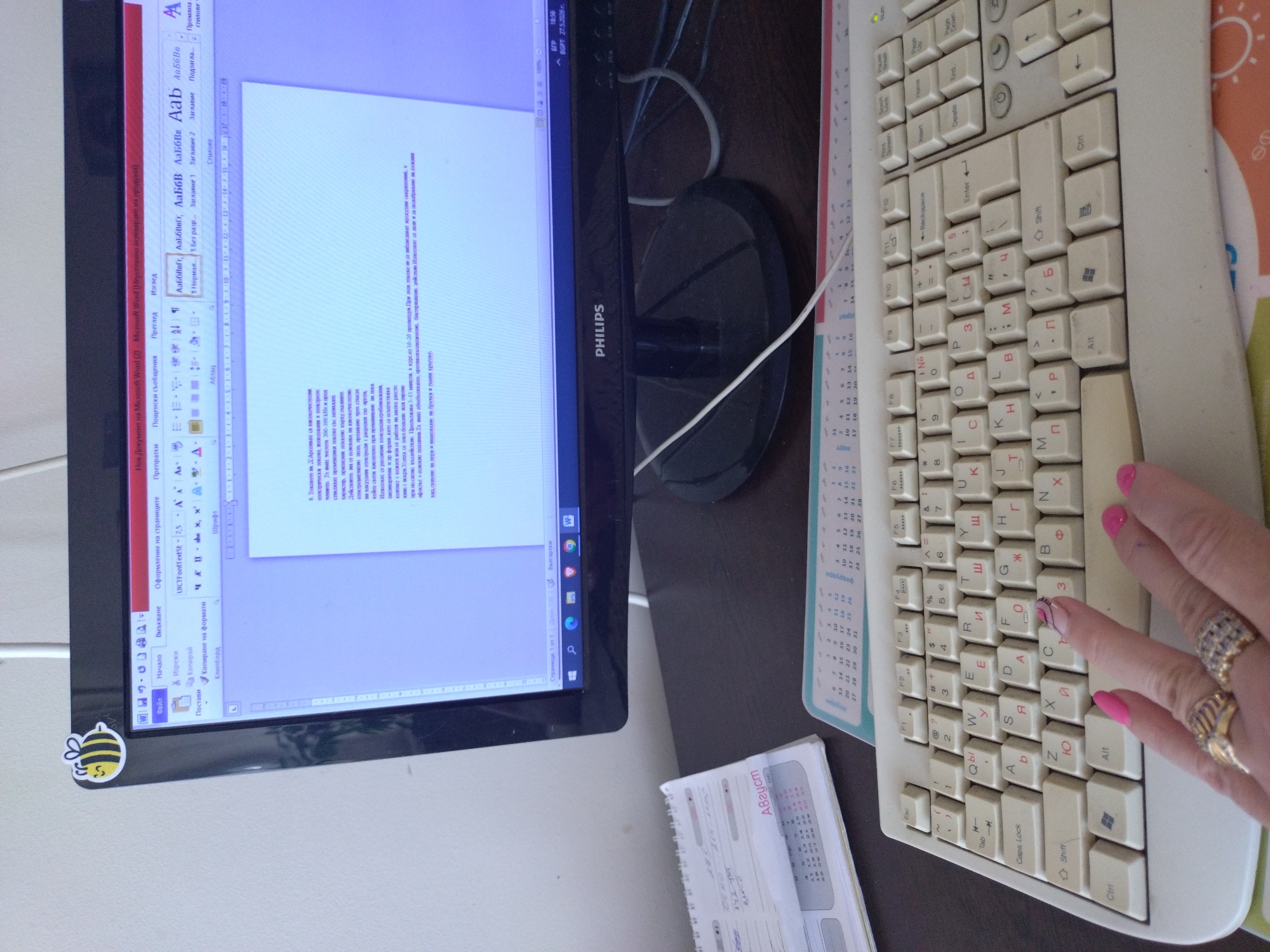 A medium, eye-level shot shows a desk setup in Unknown, Unknown, with a Philips monitor displaying a Microsoft Word document in Cyrillic, an old-fashioned keyboard with Cyrillic characters, and a small spiral notebook calendar. The monitor sits slightly to the left, displaying a text document with formatting options visible at the top. A small bee sticker is attached to the lower left corner of the monitor. To the right, the keyboard is oriented diagonally. A person's hand with manicured nails and several rings rests on the lower right corner of the keyboard. In the foreground to the lower left, a small spiral notebook calendar is visible. The desk surface has a dark brown color. The general environment appears to be a room with white walls, as seen in the background. The image is slightly rotated counter-clockwise.