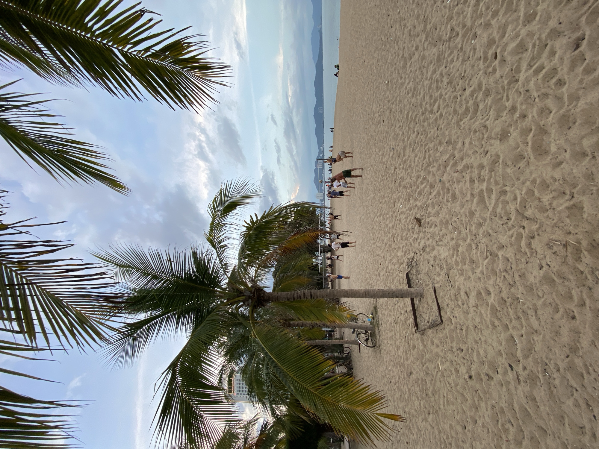 A beach scene in Nha Trang, Vietnam, is captured at dusk. The foreground is dominated by soft, textured sand, with a few palm trees casting shadows. In the mid-ground, a group of people are playing volleyball on the sand, near a volleyball net. A bicycle is parked on the beach near a palm tree. In the background, the ocean stretches to the horizon, with a few boats visible. A city skyline and mountains can be seen in the distance. The sky is overcast with clouds, and the lighting suggests the sun is setting or has recently set. The overall atmosphere is relaxed and recreational.