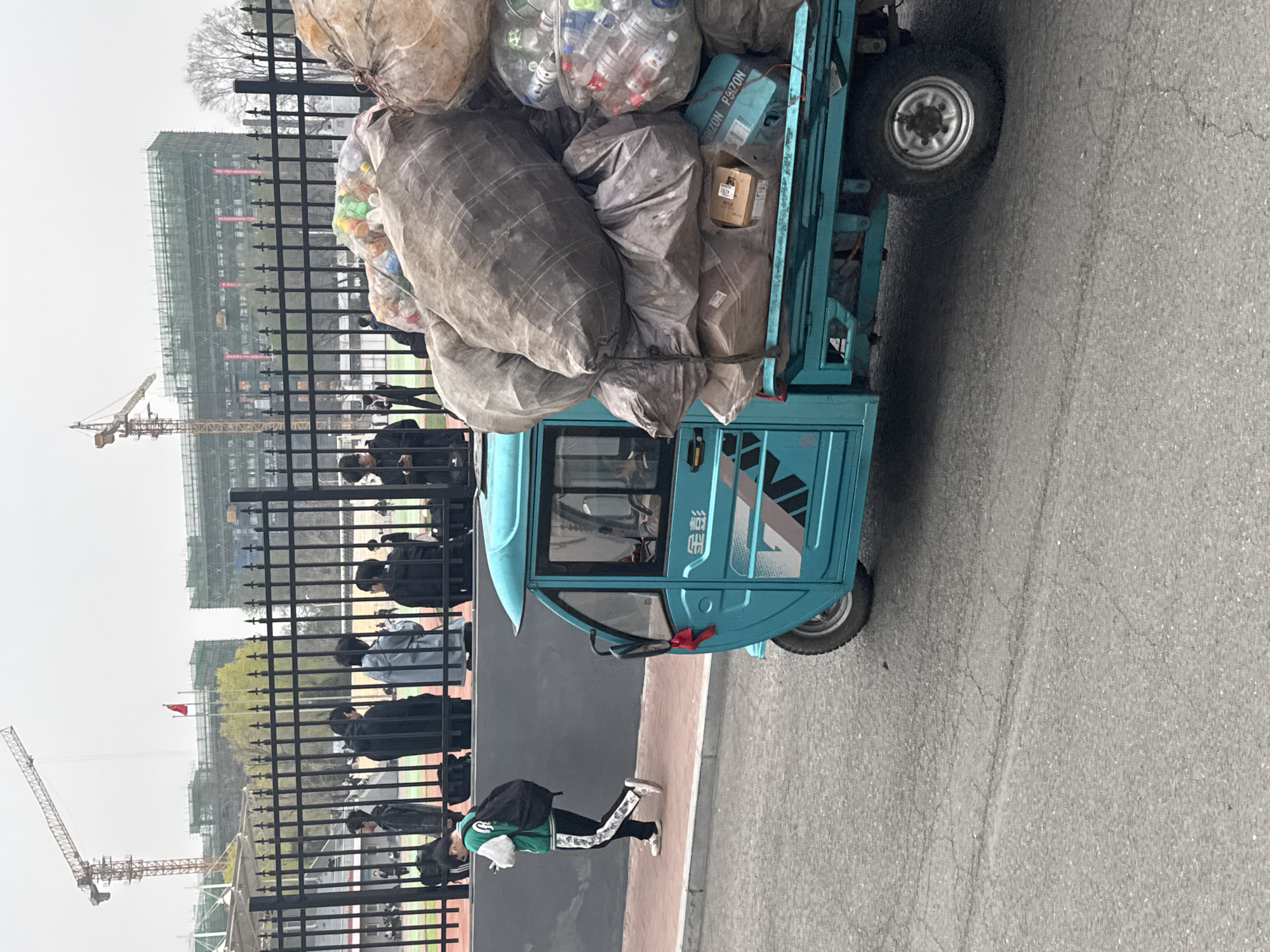 A three-wheeled blue vehicle is overloaded with bags of what appear to be recycled materials, including plastic bottles and textiles. It is parked on an asphalt road. A pedestrian wearing a green jacket and black pants with white stripes is walking on the sidewalk beside a black fence. In the background, there is a building under construction, with two cranes visible. People are standing or walking behind the fence. The sky is overcast. The vehicle appears to be an electric tricycle with a cabin. The characters on the vehicle is written in Chinese, and one of the words is 涛富. The location is presumed to be near the city of Zhongtun in China due to the prompt's instruction.