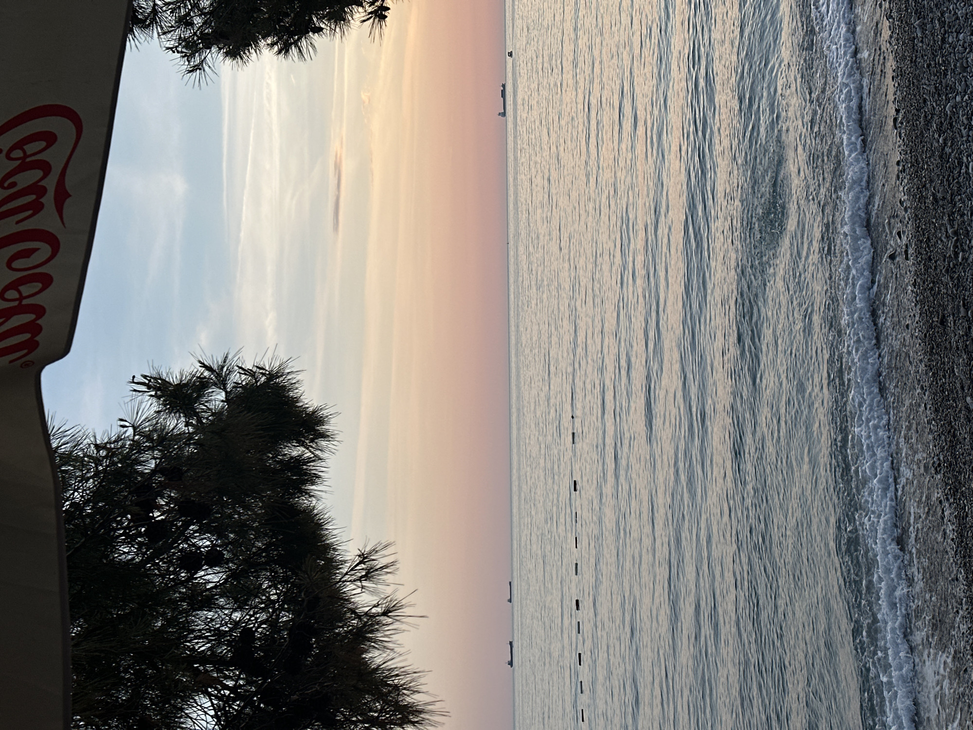 The image shows a scene at Šušanj, Montenegro, featuring a pebble beach and the Adriatic Sea at sunset or sunrise. The sky transitions from blue to pink and orange. Vegetation, including pine trees, is visible in the foreground on the left. A Coca-Cola sign is partially visible in the top left corner. In the sea, there is a faint outline of what seems to be a ship. Buoys can be seen in the sea.