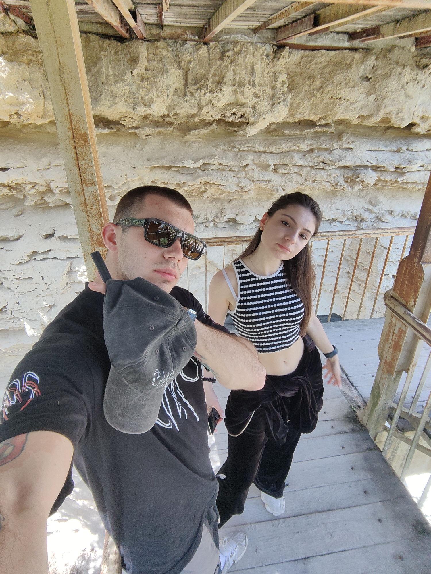A selfie photograph taken in Varna, Bulgaria, shows a man and a woman standing on a wooden platform. The man, positioned on the left, has a shaved head, wears sunglasses and a black t-shirt, and holds a black baseball cap in his hand. He has tattoos on his left arm. The woman, standing slightly behind the man to the right, has long brown hair and wears a black and white striped crop top and black velvet pants. The background consists of a textured, light-colored rock wall, wooden beams, and part of a rusty metal structure. The platform appears to be part of an elevated walkway or observation point.