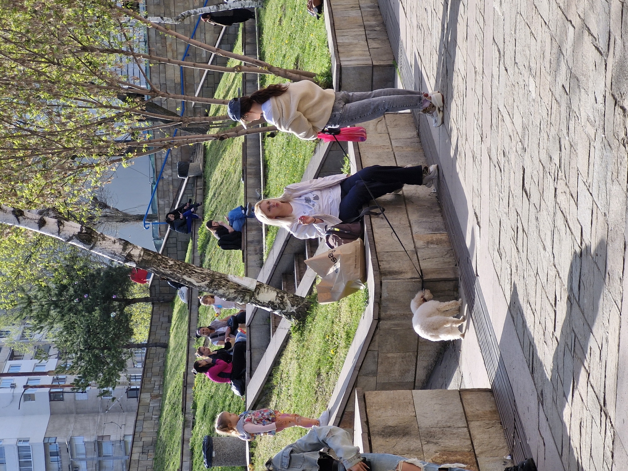 This image captures a vibrant outdoor scene in an urban park or plaza in Sofia, Bulgaria, on a bright, sunny day. The setting features multi-level paved stone pathways and steps, interspersed with grassy areas and numerous trees, including birches with light-colored bark and other trees displaying fresh spring foliage.
Many people of various ages are present, relaxing and socializing. In the mid-ground, a woman with blonde hair is seated on a paved step, holding a leash attached to a fluffy white dog that stands attentively beside her. Nearby, another woman, wearing a light-colored hoodie, jeans, and a baseball cap, stands and appears to be interacting with the dog or its owner. A red water bottle is visible attached to her clothing.
Further into the background, several groups of people are seated on the grassy slopes or steps, enjoying the sunshine. One child is seen standing upright, and colorful bubbles are floating in the air, suggesting playful activity. In the distant background, several multi-story buildings line the horizon, characteristic of an urban environment.
The strong sunlight creates clear, elongated shadows from the trees and people, indicating a daytime scene, likely morning or late afternoon. The overall mood is relaxed and social, with people enjoying leisure time in the park. A brown paper bag is placed on one of the steps near the seated woman.