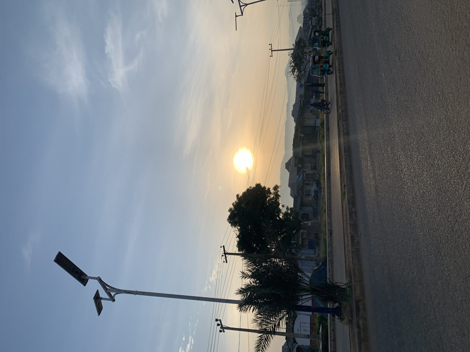 The image captures a street scene in Rafin Par, Nigeria, during what appears to be sunrise or sunset, given the low angle of the sun and the warm light illuminating the sky. The foreground is dominated by an asphalt road with a textured surface. To the left, a curb separates the road from a paved walkway. On the walkway, a solitary street lamp with a solar panel is prominently positioned. Adjacent to the street lamp is a palm tree with its fronds fanning out. Further back, several power poles with wires crisscross the scene, and a larger tree with dense foliage stands. Beyond the trees and poles, a row of low-rise buildings, likely shops or residential structures, lines the street. The architecture suggests a developing urban or semi-urban environment. Several vehicles are visible on the road: a motorcycle with a rider, two auto-rickshaws (also known as Keke Napep), and possibly another vehicle in the background. A few individuals are standing near the buildings and the auto-rickshaws, appearing to be engaged in daily activities. In the distance, a hazy outline of hills or mountains can be seen under the softly lit sky. The overall atmosphere is tranquil, with the natural light creating a peaceful ambiance over the bustling street.