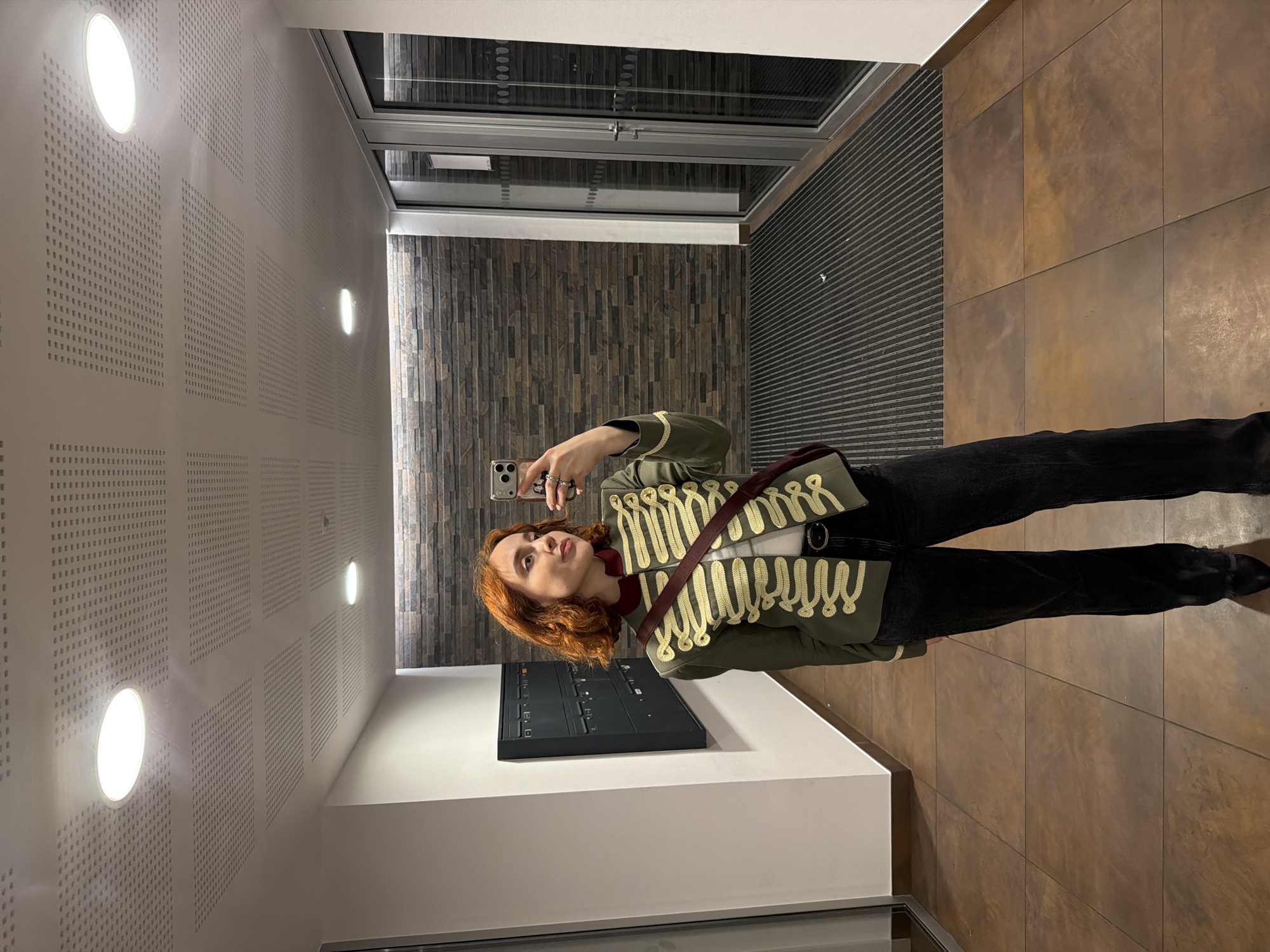 A young woman with short, reddish-brown hair is taking a selfie in a hallway. She is wearing a military-style green jacket with gold braiding, a light-colored top, black jeans, and black shoes. A dark red crossbody bag is slung over her shoulder. The hallway has a modern aesthetic with tiled floors in earthy tones, a textured stone wall, and a perforated acoustic ceiling with recessed circular lights. To her left are a set of dark gray mailboxes mounted on a white wall. To her right are glass double doors with silver frames. The scene appears to be indoors, possibly in a residential building or a public space with a contemporary design. There is no indication this location is in Strasbourg, France.