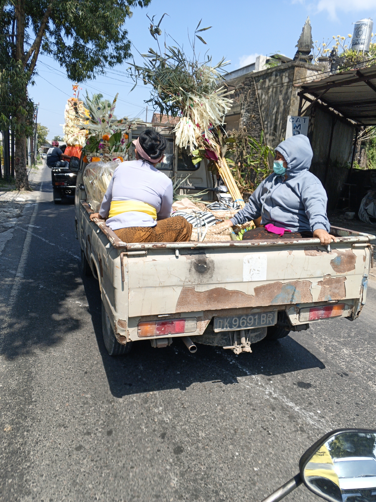 In Sukawati, Indonesia, on a sunlit day, a light-colored, weathered pickup truck drives on a paved road. The truck bed is filled with offerings and decorations, likely for a religious ceremony. Two individuals are seated in the truck bed. On the left, a person with dark hair tied back wears a light purple shirt and a yellow sash around their waist, with an orange and brown patterned skirt. On the right, a person wearing a blue hooded jacket and a light blue surgical mask is seated. The truck has a license plate that reads "DK 9691 BR". In the background, there are buildings, trees, and a clear blue sky. The road is lined with a tree on the left and a sidewalk or path on the right, with more buildings and vegetation beyond. The scene appears to capture a moment of transport related to a cultural event in Sukawati.