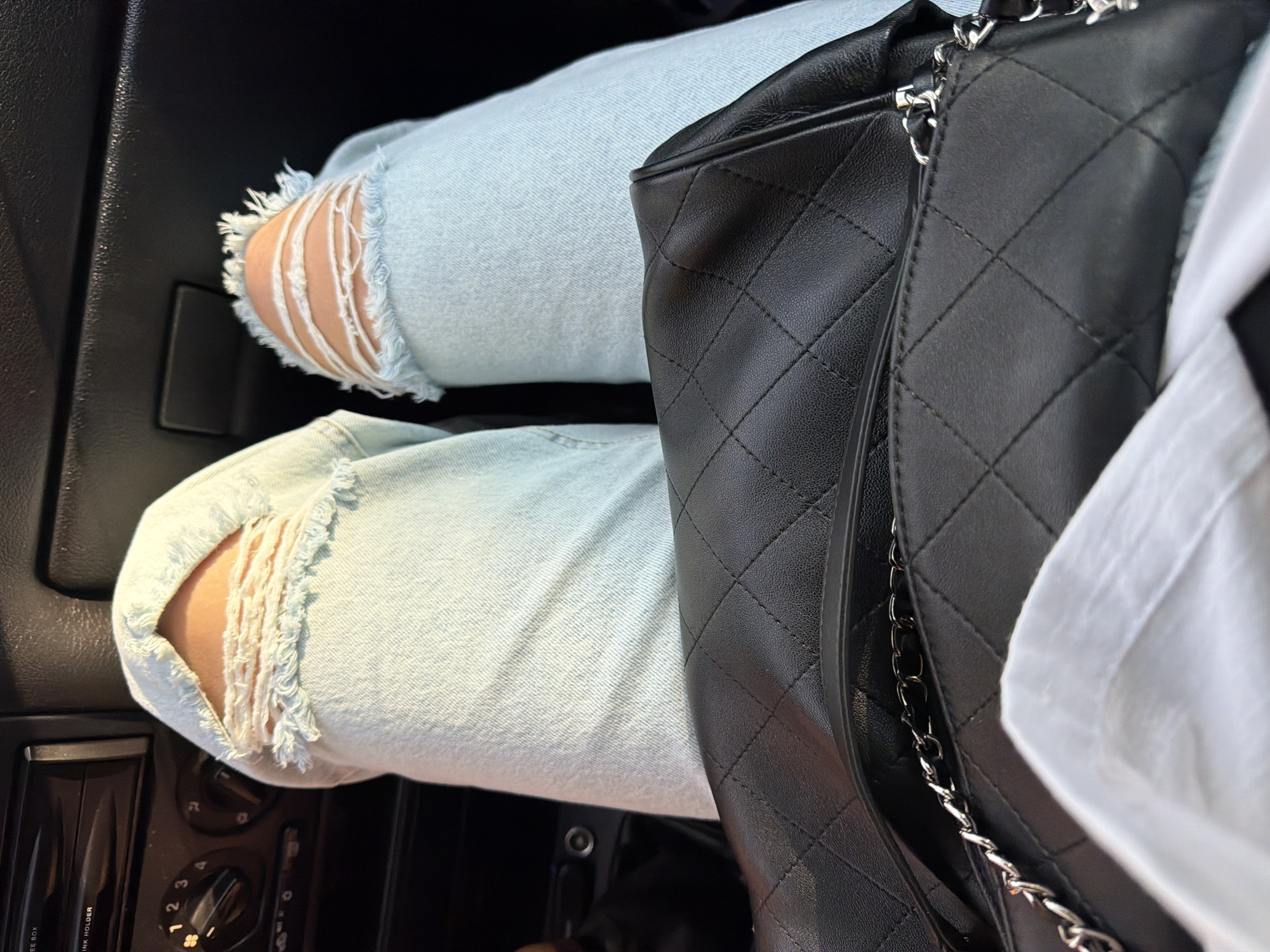 The image shows a close-up view of a person's legs inside a vehicle. The person is wearing light blue ripped jeans. A black quilted handbag with a silver chain strap is placed next to their legs. A white shirt is partially visible on the right side of the frame. The setting appears to be the interior of a car, with dark plastic elements of the dashboard visible in the background, including some dials and buttons, though no text is clearly legible. The lighting suggests it is daytime, possibly with sunlight filtering in, creating a casual and relaxed atmosphere. No specific activity is depicted other than sitting in the vehicle.