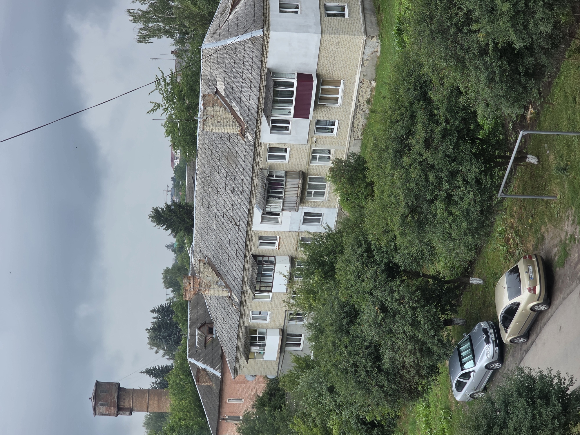 This image depicts a scene in Dubno, Ukraine, under a cloudy sky. In the background, a brick water tower stands tall, surrounded by trees. In the mid-ground, a light-colored apartment building with a gray tiled roof is visible. The building has multiple windows and balconies, with some balconies enclosed and others open. Adjacent to this building is another, partially visible, older brick building. In the foreground, two cars are parked on a dirt path next to a green, grassy area with several trees. One car is silver, and the other is gold. The overall environment is residential, with a mix of buildings and abundant greenery.