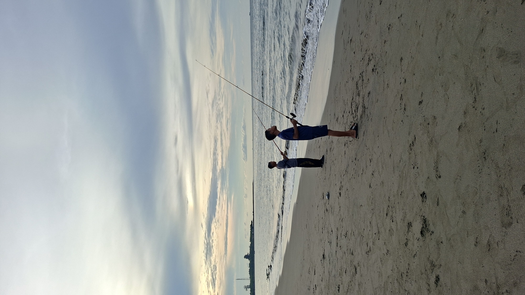 Two young boys are fishing on the sandy beach of Lhokseumawe, Indonesia, with fishing rods extended over the calm ocean. The sky is overcast with soft clouds, indicating either early morning or late afternoon. The water meets the shore in gentle waves. In the distance, a long pier or breakwater extends into the sea, with a silhouetted tower on land to the left. The sand is textured with footprints.