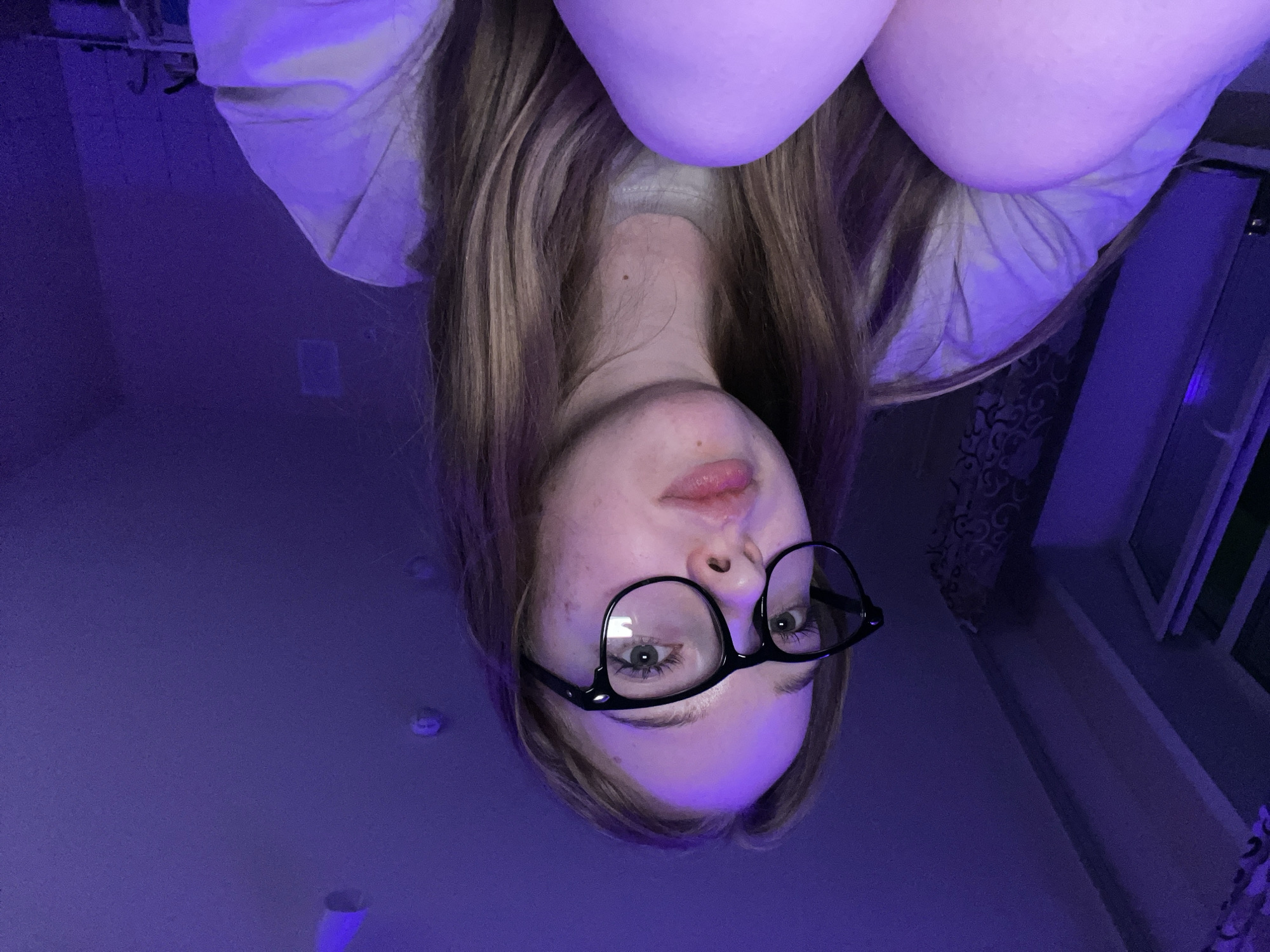 An inverted selfie depicts a fair-skinned young woman with long, light brown hair wearing black-framed glasses. She is positioned in the lower center of the frame. The lighting appears to be predominantly purple, creating a strong color cast on the scene. Behind her are indistinct details of a room, including a window and a wall with a patterned covering. The location is difficult to determine from the visual data. However, the city of Chaikovsky, Russia, cannot be confirmed or denied based on this information.