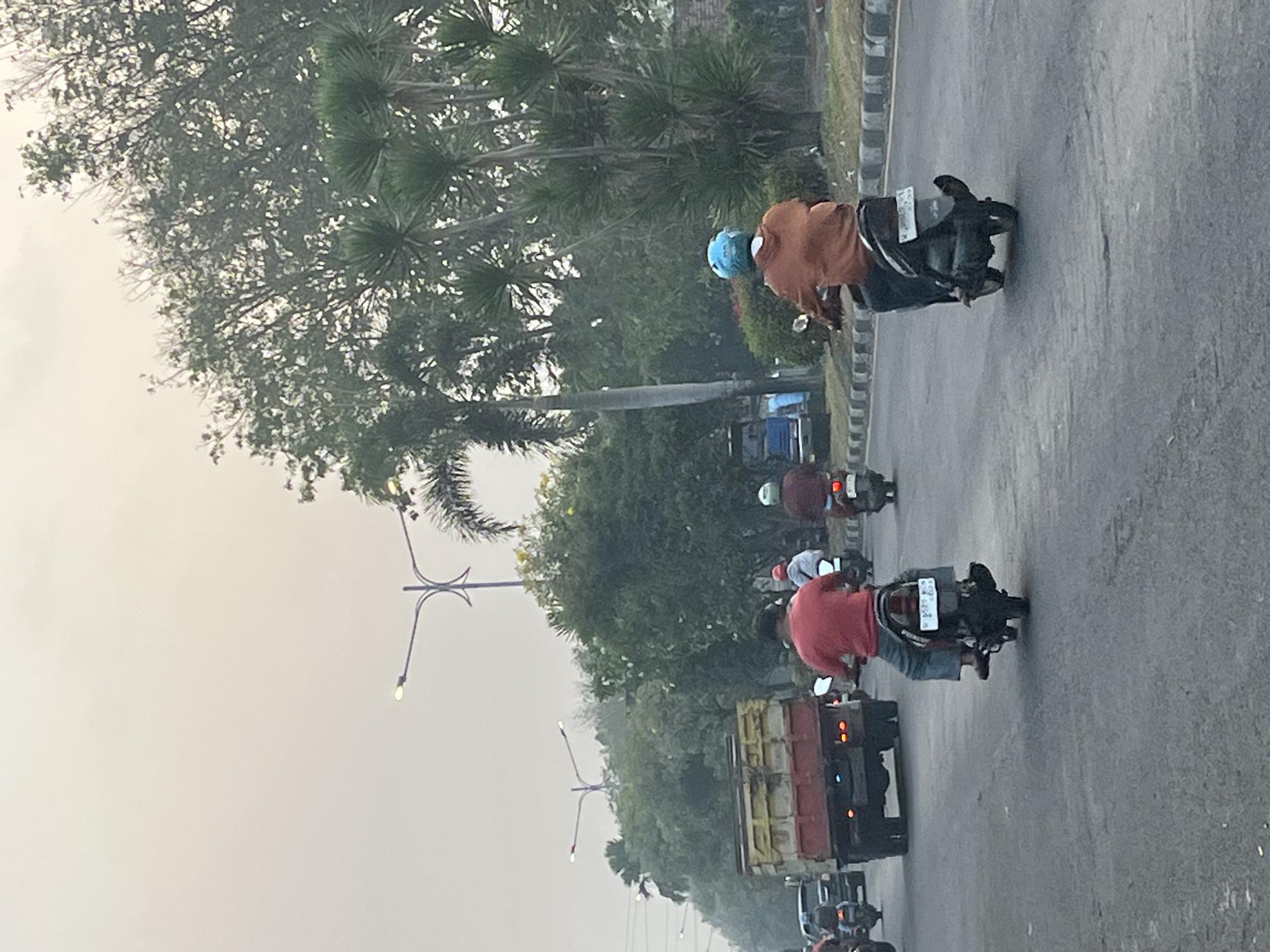 The image captures a street scene in Kludan, Indonesia, during daylight with an overcast sky. The focus is on a road with several vehicles, primarily motorcycles and a truck. In the foreground, two individuals are riding motorcycles. The rider on the left is wearing a red shirt and blue jeans. The rider on the right is wearing an orange hooded jacket and a blue helmet. Further down the road, other motorcycles and a red and yellow truck are visible. The environment includes a row of trees and palm trees lining the road, with streetlights and power lines also present. The road surface appears to be asphalt, with some visible cracks. The overall impression is of a moderately busy urban or suburban environment.