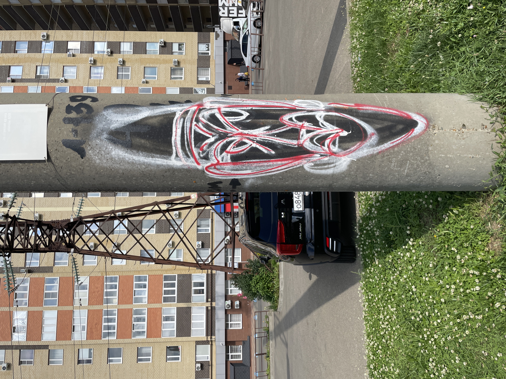 The image depicts an urban outdoor scene, likely in the city of Stavropol, Russia. In the foreground, a concrete utility pole stands prominently, adorned with graffiti. The graffiti features a stylized, abstract face drawn in white and red spray paint, with black accents. The face is elongated and has a somewhat tribal or mask-like appearance, with large eyes and a pointed chin. Above the face, the number "1-139" is scrawled in black paint.
Behind the pole, a black SUV is parked. The car's rear is visible, and it has a license plate that reads "0848 GM". To the left of the car, a patch of green grass with small white flowers is present.
In the background, a tall, multi-story brick building with numerous windows can be seen. The building has sections of light yellow and reddish-brown brick. Adjacent to the building, a large, metallic power transmission tower with intricate latticework is visible. The tower is dark brown and appears to be made of weathered steel. Further in the background, other buildings and vehicles are discernible, suggesting a residential or mixed-use urban environment. The sky is bright, indicating daytime. The overall scene is one of everyday urban life, with elements of infrastructure, vehicles, and graffiti art.