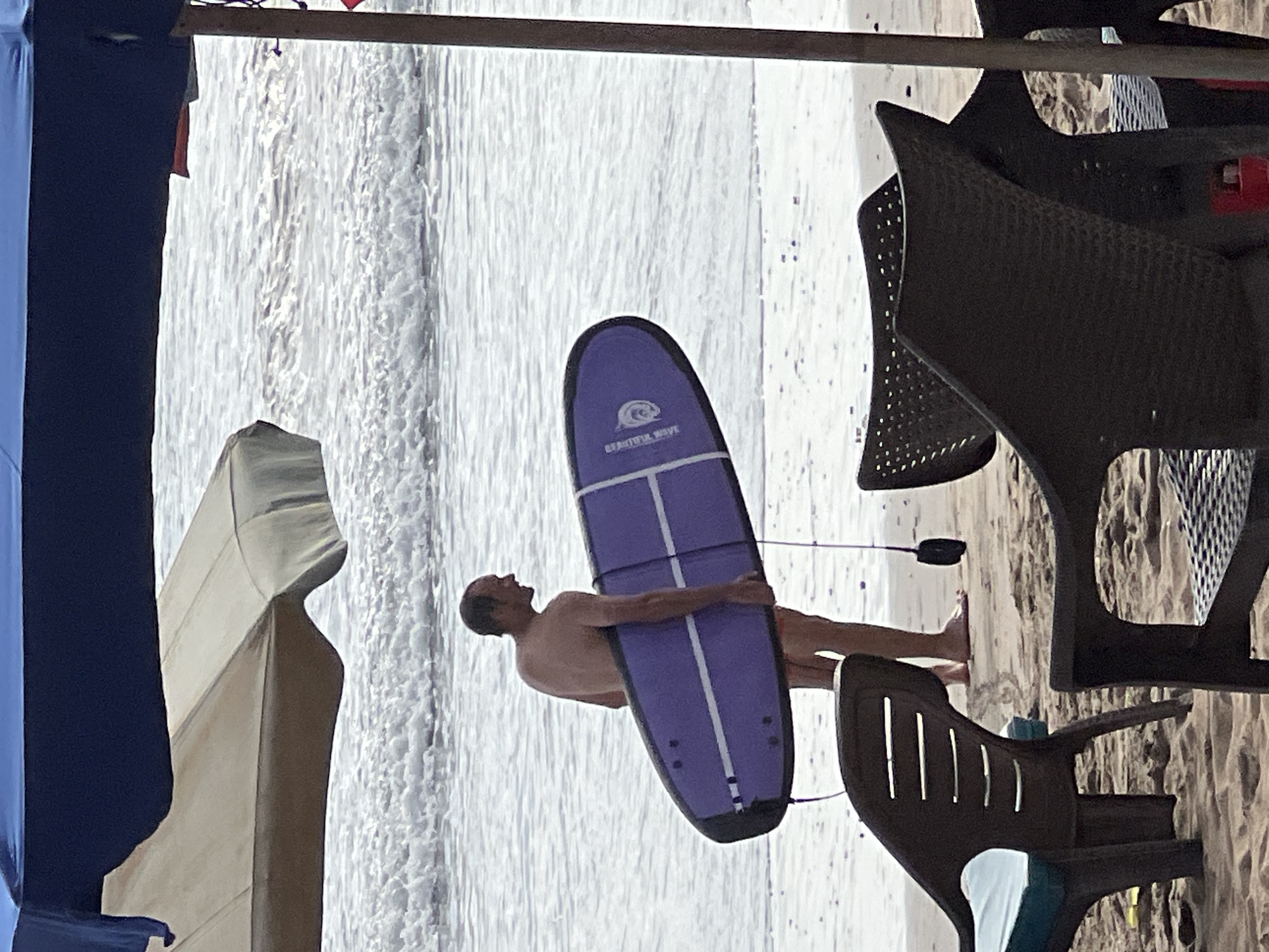 A fair-skinned man, appearing to be in his late 40s or early 50s, stands on a sandy beach, holding a purple surfboard under his right arm. He is shirtless and wearing red swim trunks. The surfboard has two white stripes running lengthwise. The man is facing to his right and appears to be looking at the ocean.
In the foreground, several dark plastic beach chairs are visible, some with cushions. The beach is covered in sand. Behind the man, the ocean waves are breaking. There are also two beach umbrellas visible, one tan and white, and one dark blue. The scene appears to be a beach in Kuta, Indonesia, given the presence of surfboards and beach umbrellas typical of the area. The lighting suggests it is daytime, possibly late afternoon or early morning, as the light is diffused.