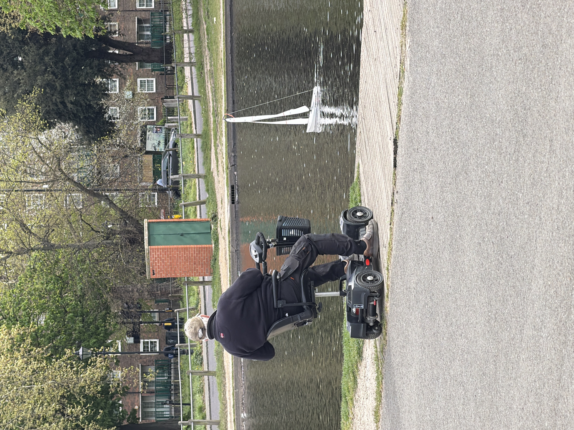 An older adult male with light skin wearing a light-colored head covering, dark jacket, and dark pants is seated in a black mobility scooter on a paved path next to a body of water. The water contains a model sailboat. On the far side of the water are green grassy areas and a footpath with a wooden railing. Behind that, there are brick buildings with multi-paned windows. A small brick and green shed is also visible. The scene is possibly in London, United Kingdom, based on the architecture.