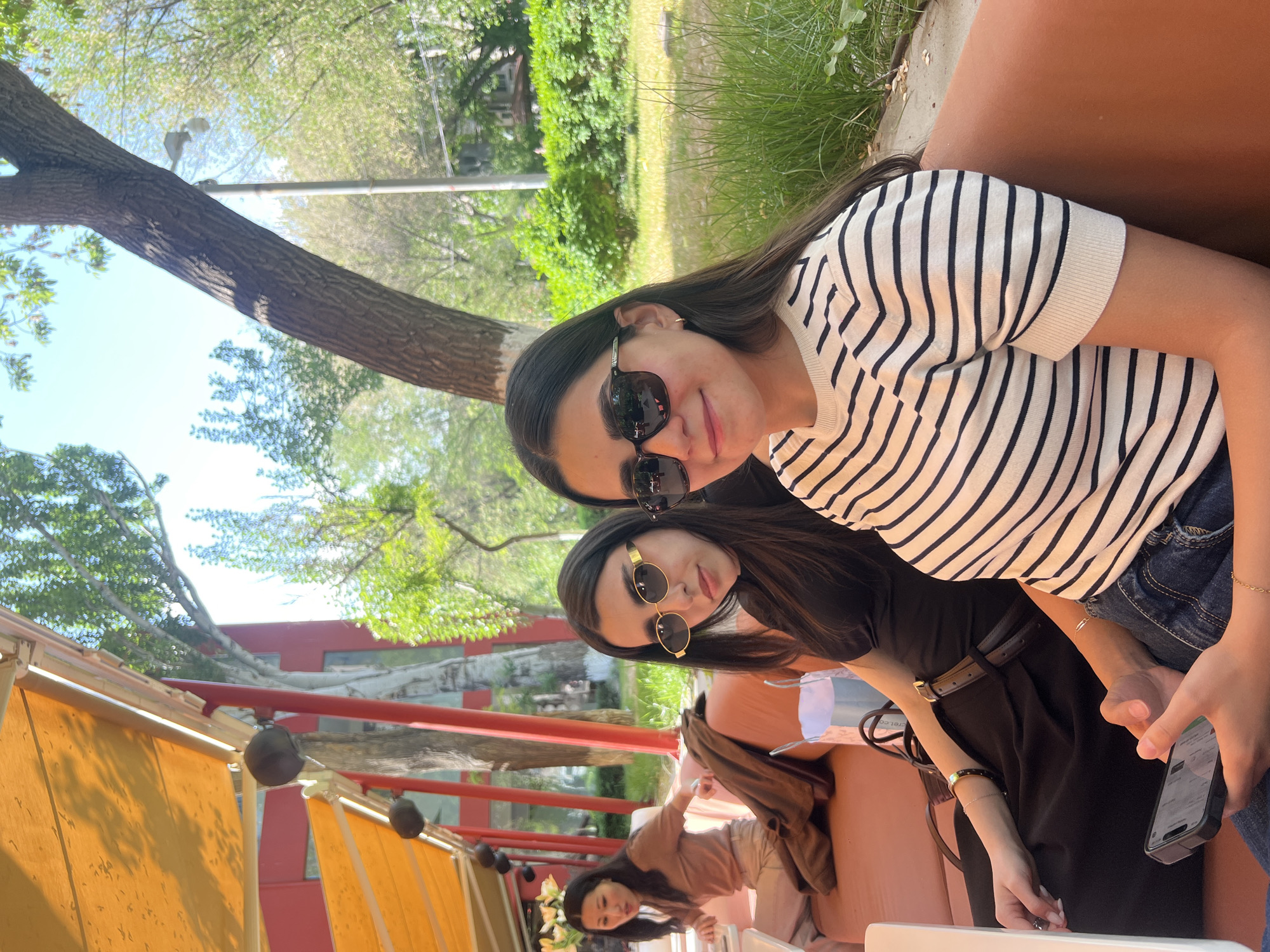The image depicts two women in sunglasses sitting close together at an outdoor cafe or restaurant. The woman in the foreground wears a striped black and white shirt, while the other wears a black dress. A third person, a woman, is seated farther away at the table. A smartphone is visible in the hand of the woman in the black dress. The environment includes trees, grass, and what appears to be an awning with light orbs above the tables. The location appears to be in the city of Unknown, Unknown.