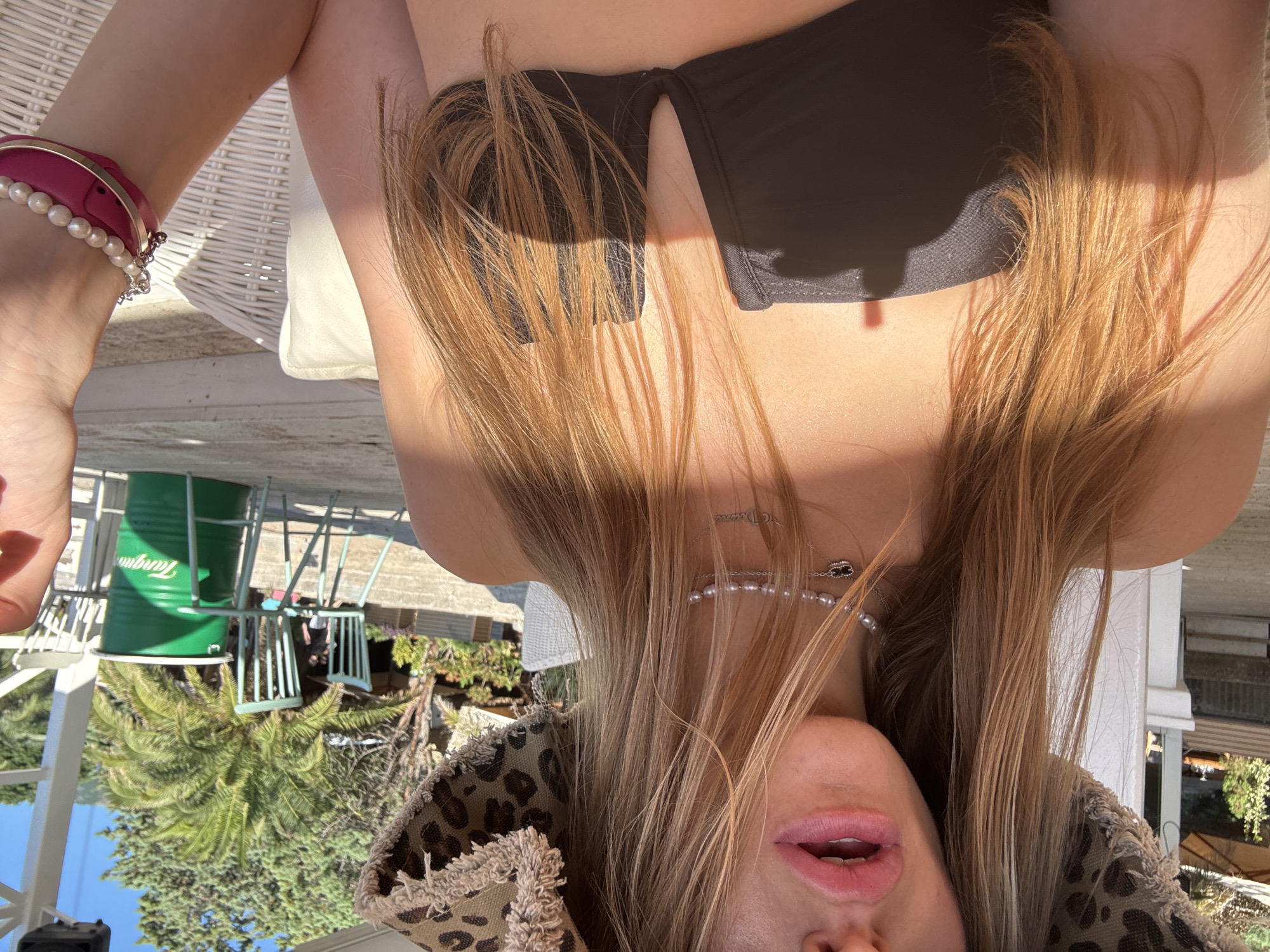 A close-up, over-the-shoulder, selfie-style shot shows a person wearing a leopard-print bucket hat and a black bikini top. The person's mouth is slightly open, and they are wearing a pearl necklace and a pendant. Their hair is long and blonde. The background appears to be an outdoor setting with palm trees, chairs, and a green barrel. The location is identified as Kamenovo, Montenegro.