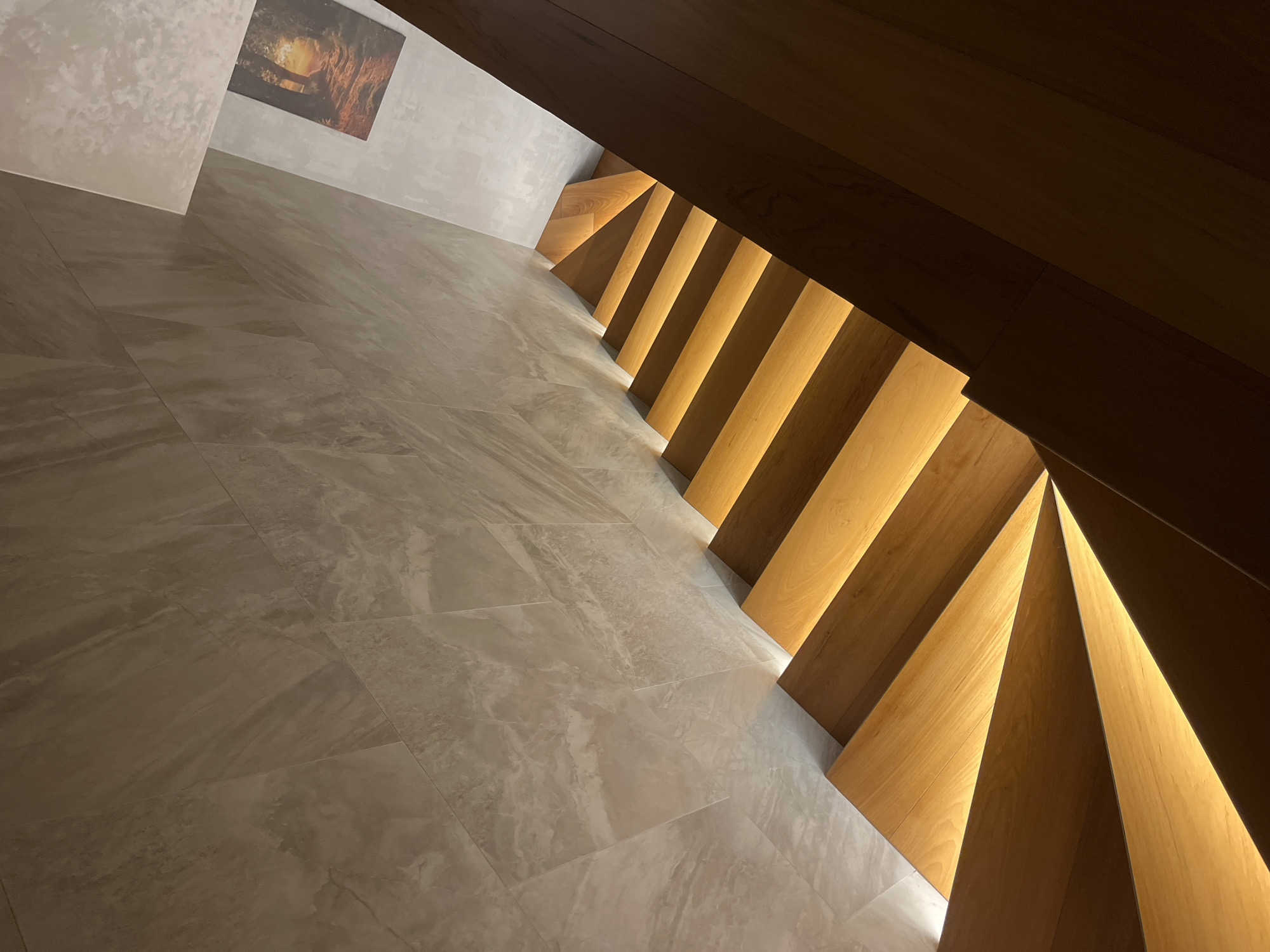 The image depicts an interior scene with a focus on a staircase and flooring. The staircase is constructed with wooden steps, each appearing to have integrated lighting that illuminates the gaps between the steps, creating a pattern of light and shadow. The steps are arranged in a staggered pattern, adding a modern or artistic design element. The flooring consists of large, rectangular tiles with a neutral, stone-like appearance. These tiles cover a significant portion of the floor.
In the upper left corner, there is a glimpse of a wall with a textured finish, and a framed artwork is visible, depicting what seems to be a fiery or cave-like landscape. The overall color palette is warm, with tones of beige, brown, and yellow. The lighting is a combination of natural light entering from an unseen source and the artificial lighting from the staircase. The scene is architectural in nature and does not contain any visible people. There is no information available in the image about the location, city, or country.