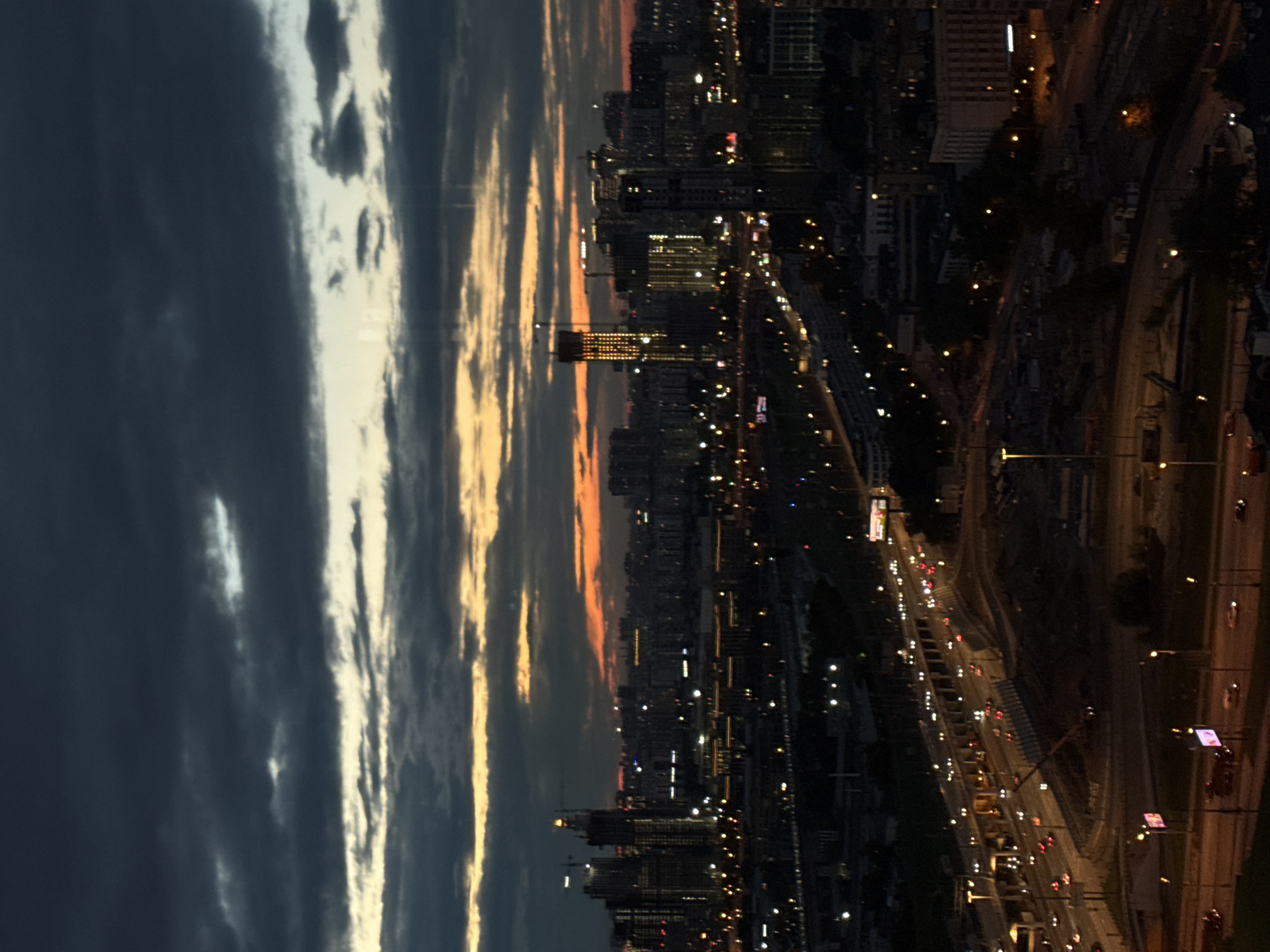 This is an aerial, wide-angle photograph of the city of Khimki, Russia, taken at dusk or dawn. The sky is overcast with dark, dramatic clouds, with bands of orange and yellow light from the setting or rising sun visible near the horizon. The city skyline is dominated by numerous high-rise buildings, including several under construction, some of which are illuminated. A multi-lane highway with moving vehicles and streetlights is prominent in the foreground, curving through the urban landscape. Construction cranes are visible among the buildings. The overall scene depicts a developing urban environment under a moody sky.