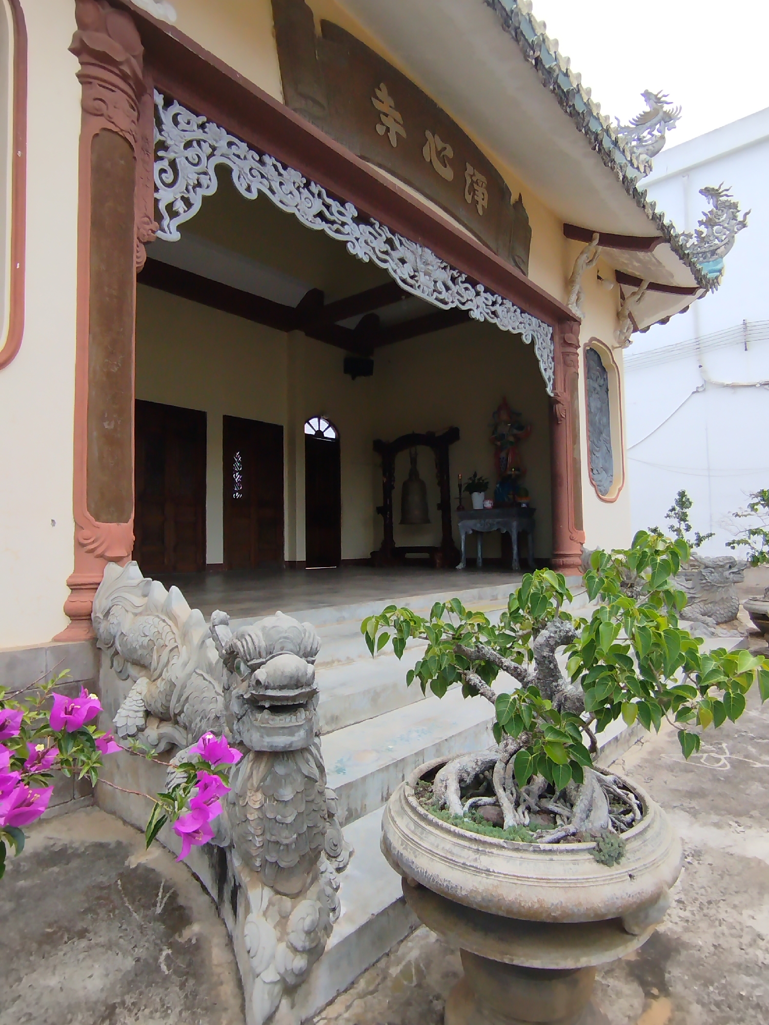 The image shows a temple-like building in Phú Lộc, Vietnam. The building has a light yellow facade with brown and white ornamental details. A sign with Asian characters is visible above the entrance. Stone dragon statues flank the steps leading up to the entrance. A bonsai tree in a large pot is in the foreground. Inside the building, a bell and altar with a statue are visible. The scene is captured during the daytime, and the environment appears serene and well-maintained. The architecture indicates a cultural or religious site.