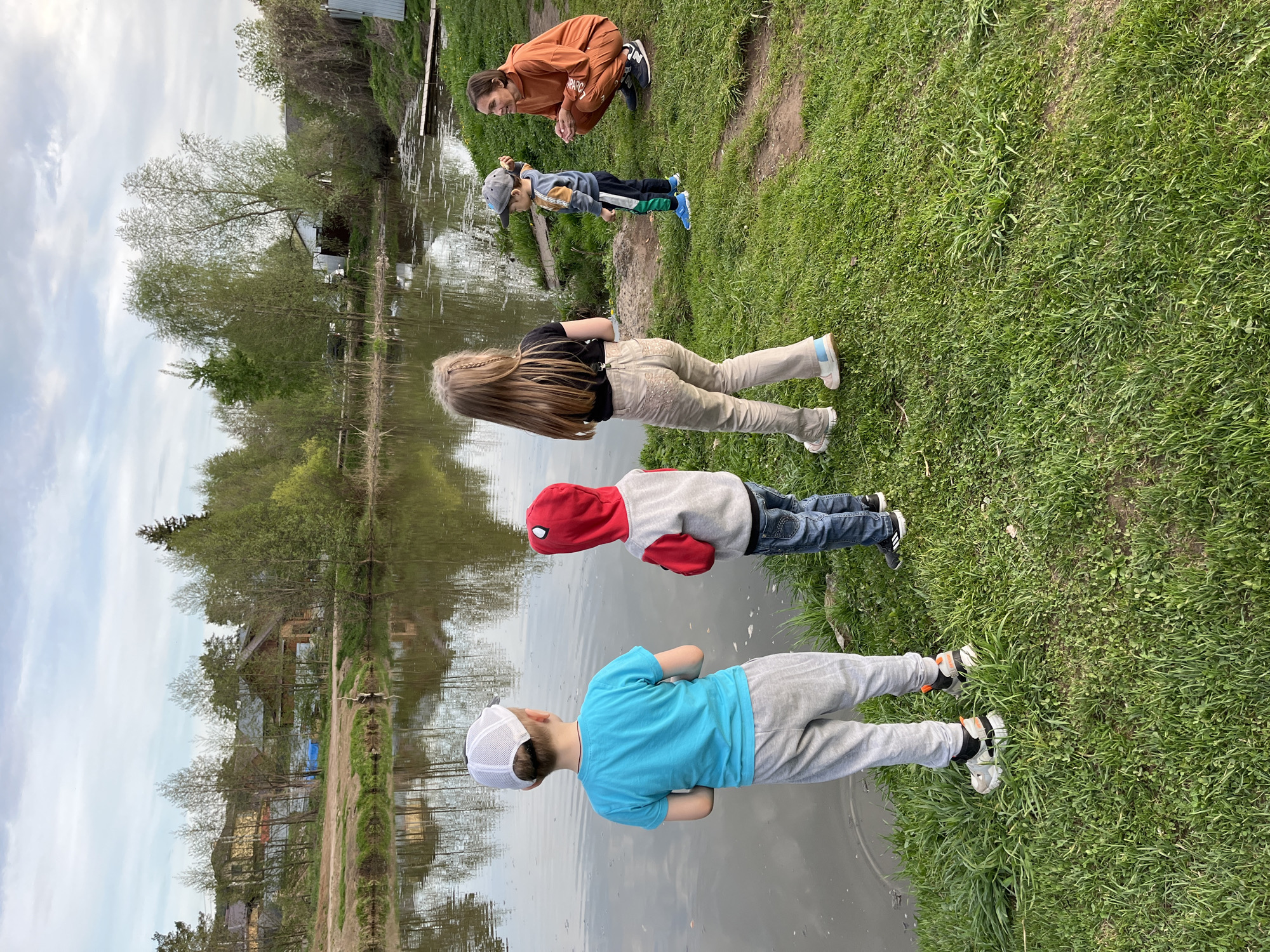 The image shows five people near a body of water. The location appears to be a grassy area next to a pond or lake, possibly in a rural or suburban setting with visible houses and trees in the background. From the angle the photo was taken it is difficult to say anything else regarding the location. Five people are standing near the water's edge. One individual is crouching, and the other four are standing. The individuals are wearing casual clothing. There appears to be a possible interaction between the individuals, however the exact nature of the interaction is indiscernible.