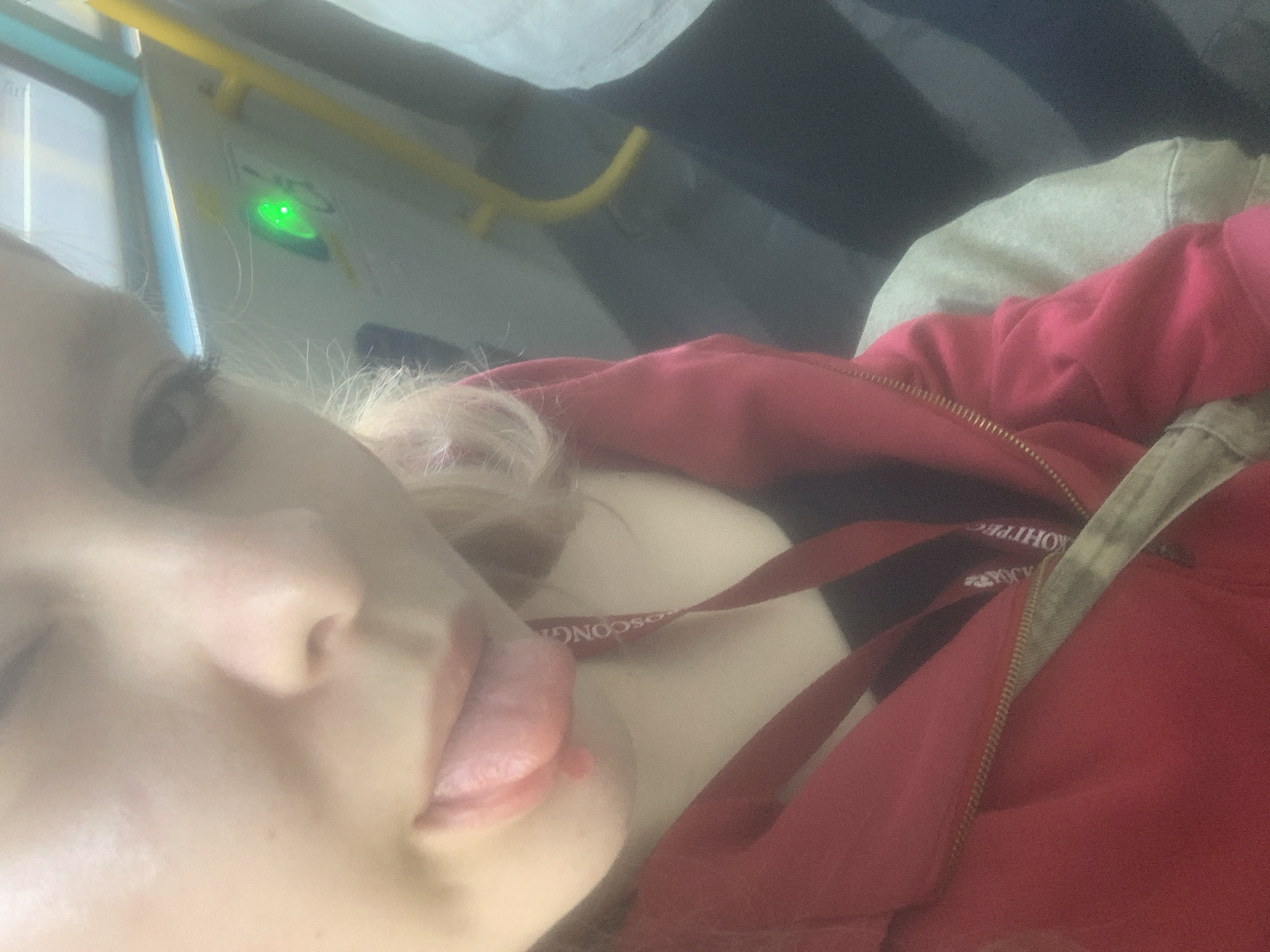 The image is a close-up, slightly blurry selfie taken from an overhead perspective, depicting a person from the chin upwards. The individual has blonde hair and is seen sticking their tongue out, suggesting a playful or casual mood. They are wearing a red zippered hoodie or jacket over a lighter-colored top.
The background elements indicate an interior setting, likely within a public transport vehicle such as a bus. A bright yellow handrail extends across the upper portion of the frame. Below it, a distinct green light glows on a light-colored panel, possibly indicating a system status or a stop request button.
A red strap or ribbon, potentially from a lanyard, is visible across the person's chest. Mirrored text on this strap reads "МОСКОНГ..." and "...ОНЦЕРТ", strongly suggesting the word "МОСКОНЦЕРТ" (Mosconcert), an organization based in Moscow. The image is taken indoors, with diffused lighting, and shows no specific time of day or weather conditions.