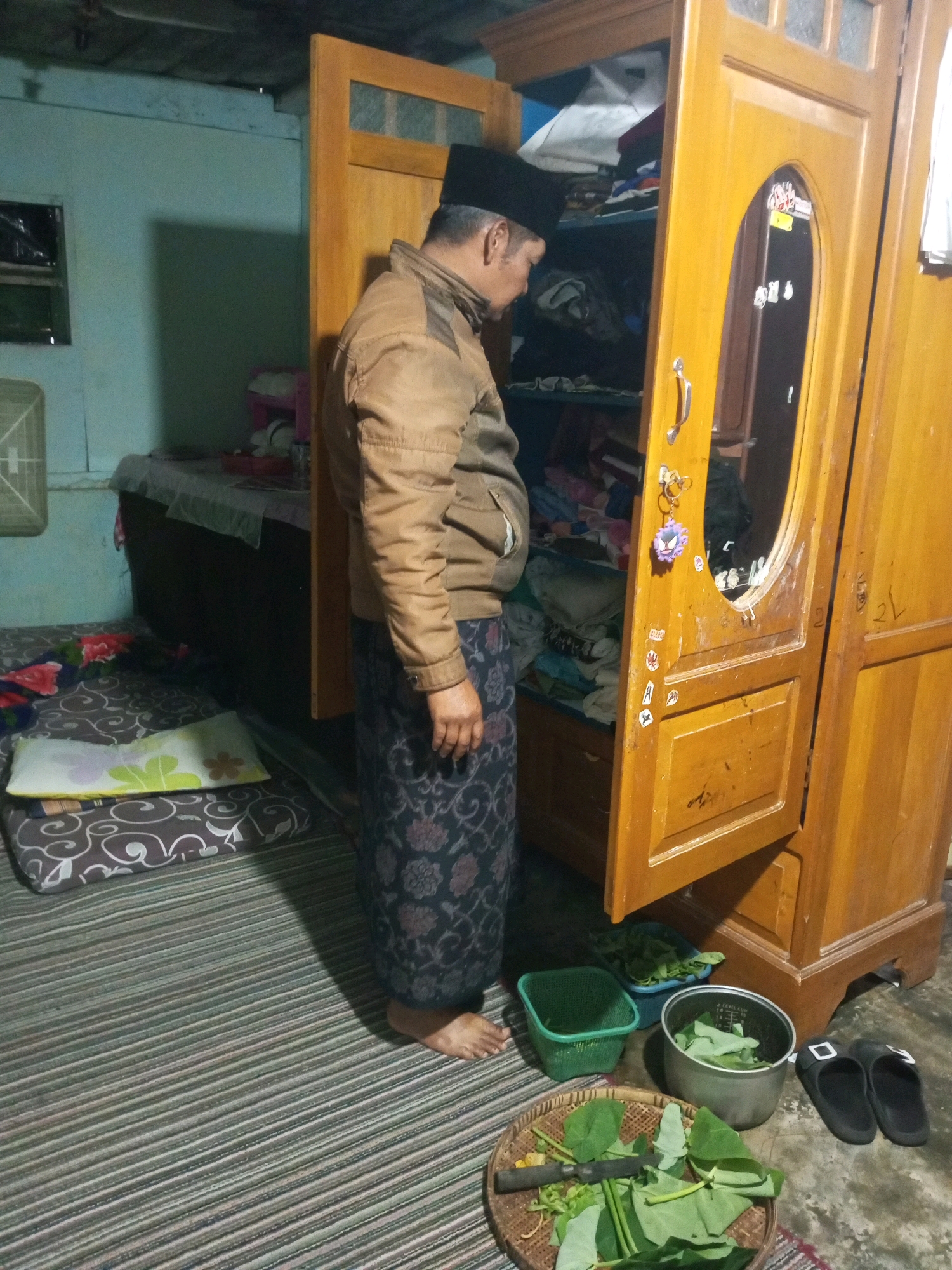 In a dimly lit, modest room, likely in Andongsili, Indonesia, a person wearing a black peci cap, a brown jacket, and a patterned sarong stands facing an open wooden wardrobe. The wardrobe is filled with clothing and other items. In front of the wardrobe, on a striped mat, are several objects: a green plastic basket, a grey pot filled with green leaves, a woven tray with more green leaves and a knife, and a pair of black slippers. A mattress with a patterned cover and a pillow is visible on the left side of the image. The walls of the room are painted a light blue, and there is a window covered with plastic on the left wall. The overall impression is one of a domestic, utilitarian space.