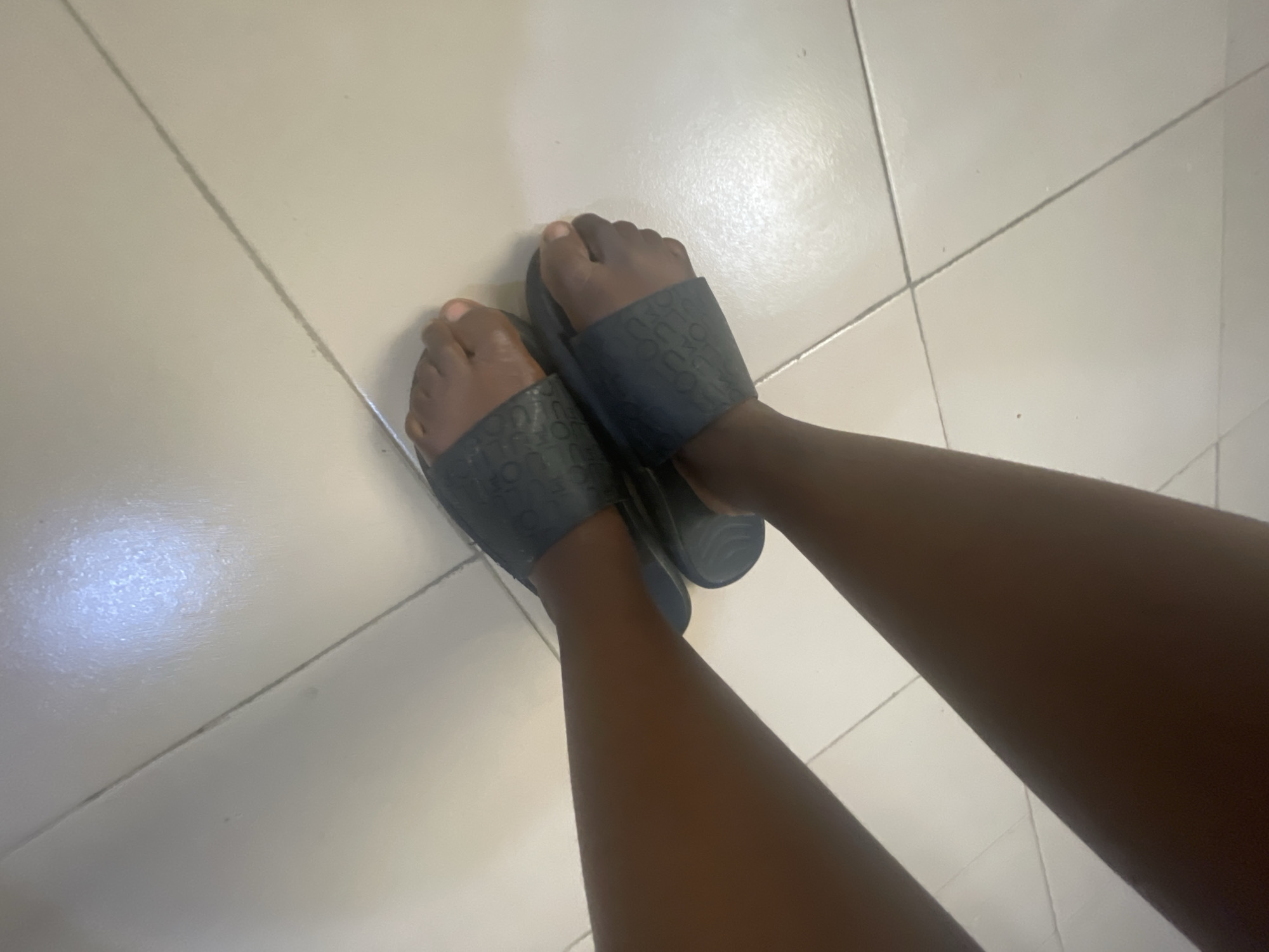 This media file shows a close-up, overhead view of a person's feet wearing dark-colored slide sandals. The sandals have a textured pattern on the straps, possibly a repeating logo. The person's skin tone is dark. The feet are positioned on a tiled floor with visible grout lines. The tiles appear to be a light, neutral color, possibly beige or off-white. The lighting suggests it might be daytime, as there is a noticeable reflection or glare on the tiles, indicating an overhead light source or natural light. The scene appears to be indoors, given the tiled flooring. The overall impression is casual and domestic. There is no visible text in the image. The location context is Jalingo, Nigeria.