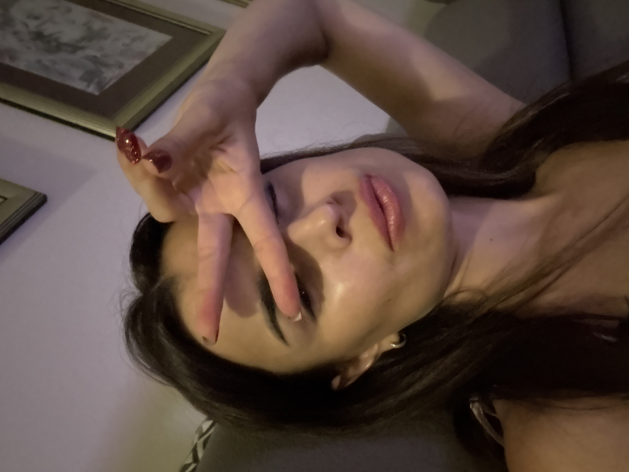 A young woman with long, dark hair is holding her hand up in front of her face, making a peace sign. She is wearing lip gloss and has hoop earrings. Her nails are painted a dark red. The background shows a wall with framed pictures. The lighting is dim, suggesting an indoor setting at night.