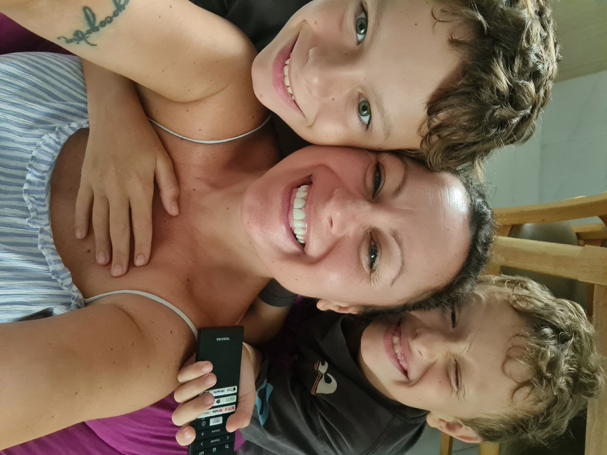 A woman is lying down, holding a black TOSHIBA TV remote in her left hand, with two boys smiling and positioned above and below her, their faces close to hers. The woman has dark hair pulled back and a wide smile. The boy above has curly brown hair and green eyes. The boy below has blond, curly hair. A tattoo is visible on the woman's left arm. She is wearing a white and blue striped top. The boys are wearing grey T-shirts. The background is slightly blurred, suggesting a room. The image does not provide any information about the location in the city of Đà Nẵng, Vietnam.