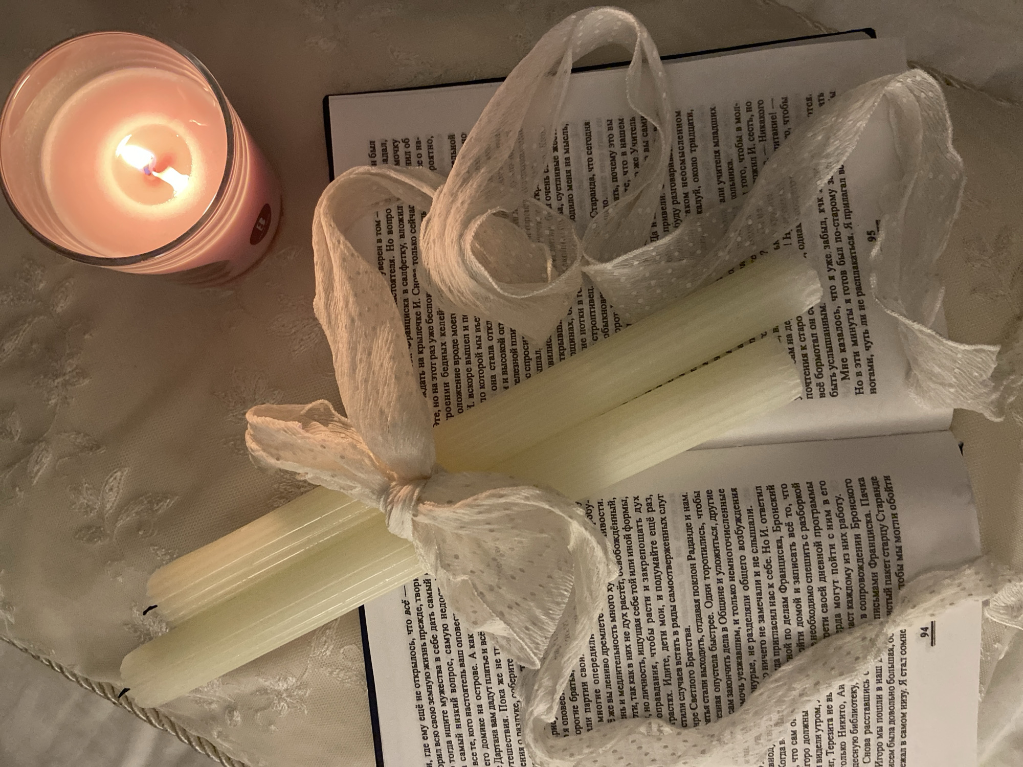The image shows a book lying open on a light-colored patterned surface, with two long, white candles tied together with a delicate white ribbon. A pink candle in a glass holder is burning in the background. The book’s pages are filled with text in what appears to be the Cyrillic alphabet. The overall setting has a soft, muted tone, creating a contemplative mood. The image does not contain sufficient detail to provide information about the location or city of Budva, Montenegro.