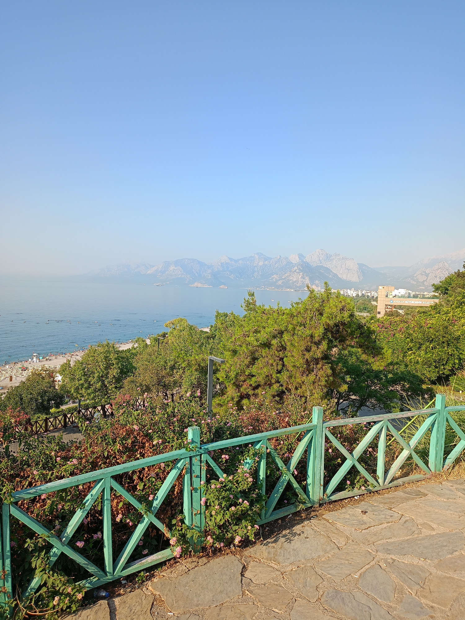 The image captures a panoramic view from a higher vantage point overlooking the coast of Antalya, Turkey. In the foreground, a weathered, light brown stone pathway leads to a bright green, wooden fence with a distinctive criss-cross pattern. This fence encloses a lush garden filled with pink flowers and green foliage. Beyond the fence and garden, a broad expanse of the Mediterranean Sea stretches out, dotted with numerous small boats and what appear to be paddleboarders. A sandy beach, populated with many sunbathers, is visible along the coastline. Further in the distance, the city of Antalya unfolds, with buildings nestled against a backdrop of majestic, rugged mountains. The sky above is a clear, pale blue, with a slight haze obscuring the furthest mountain peaks. The overall scene suggests a warm, sunny day in a coastal resort area.