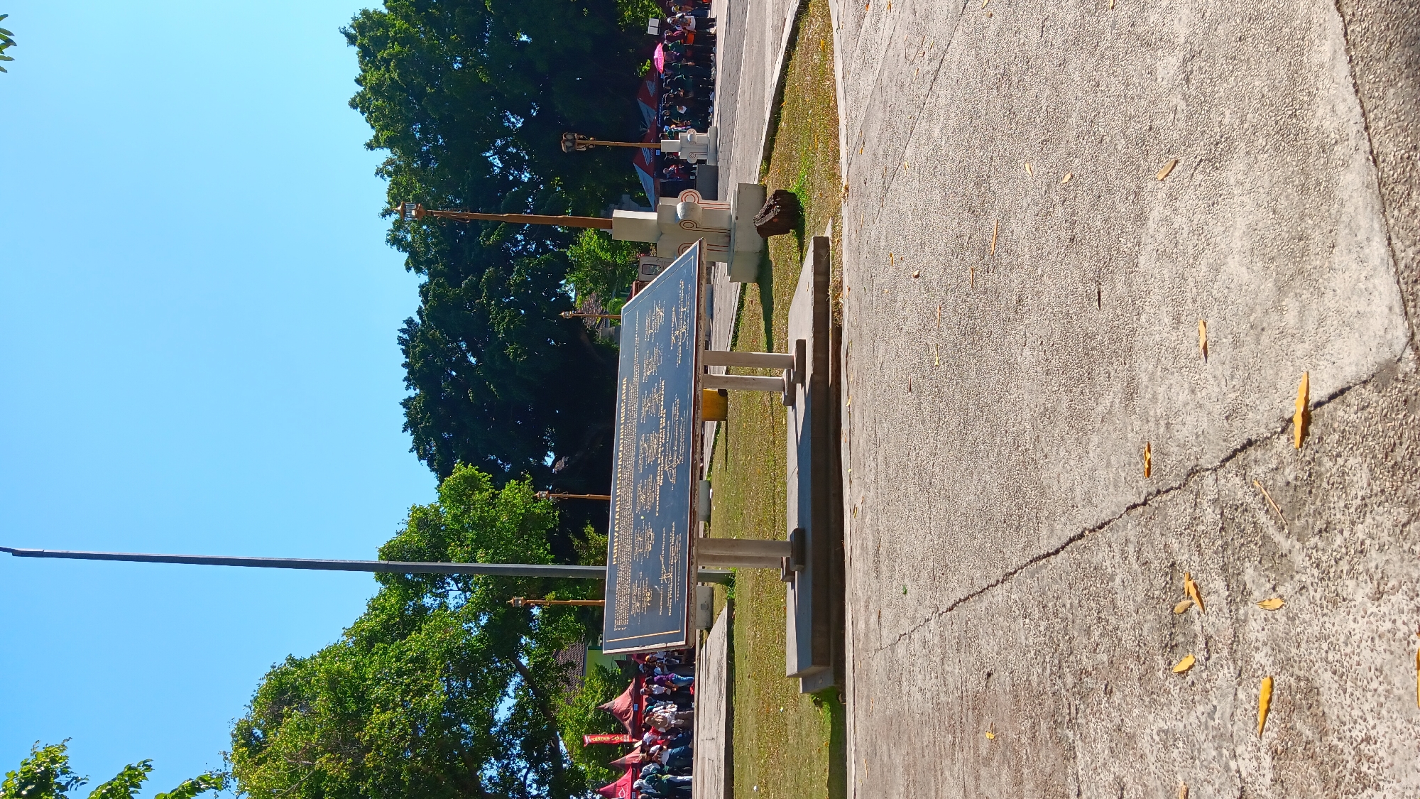 The image depicts an outdoor scene on a sunny day, likely in a public park or square, located in Nganjuk, Indonesia. In the foreground, a concrete pathway shows cracks and scattered yellow leaves. The central subject is a large, dark plaque mounted on two white pillars, situated on a raised concrete base. The plaque contains text in Indonesian, with a prominent title that appears to be "PERNYATAAN KESEPAKATAN BERSAMA" (Joint Agreement Statement).
Behind the plaque, a grassy area leads to a pathway and a road where several people are gathered. Some individuals are under red tents on the left side of the image, while others are walking on the right side, some holding umbrellas. Trees with lush green foliage surround the area, and a tall flagpole stands to the left of the plaque. Two decorative lampposts are visible further back, one to the right of the plaque and another near the gathered crowd. The overall environment suggests a public gathering or event taking place in an urban setting.