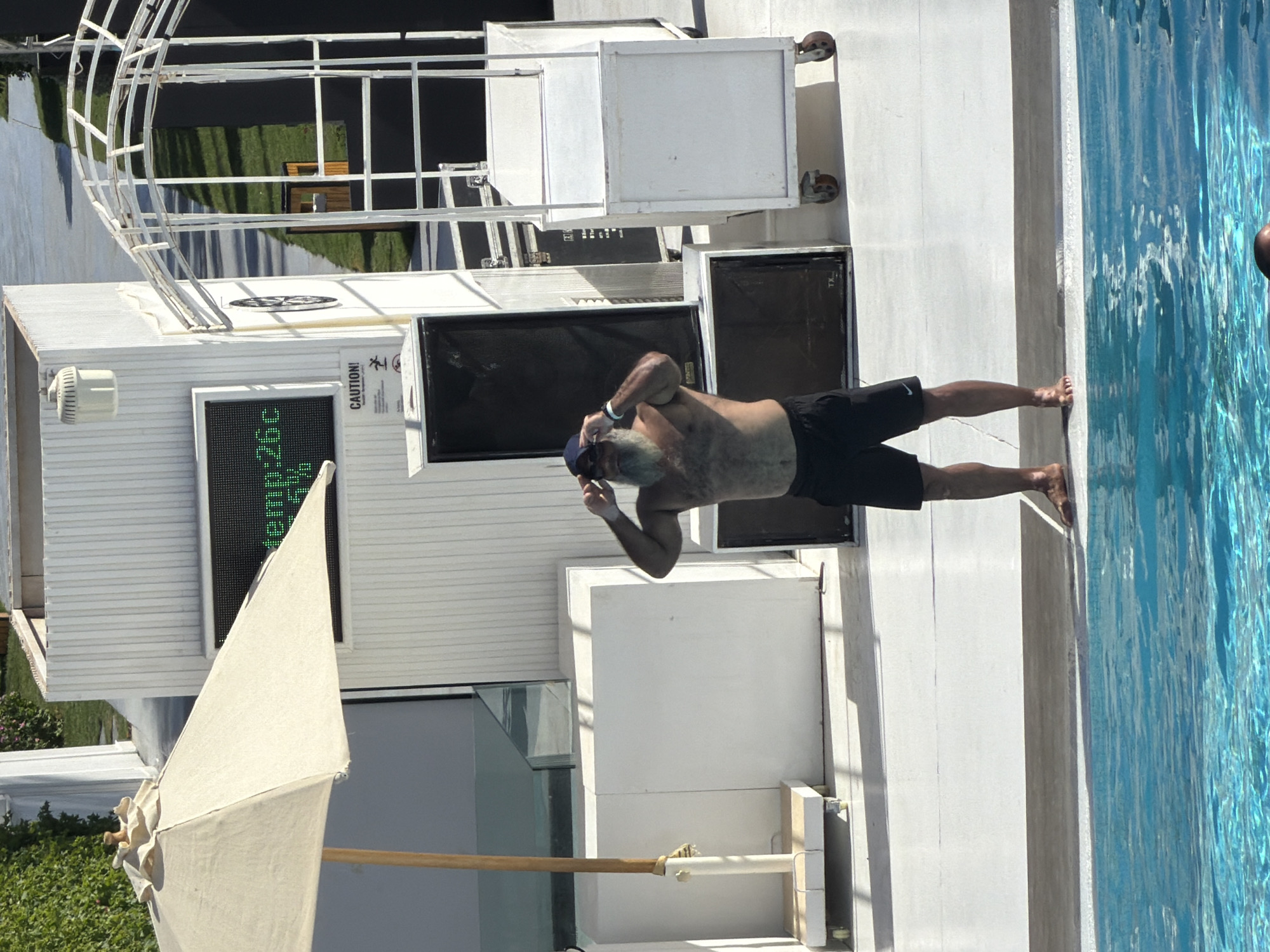 An adult male with a long grey beard, wearing a dark blue baseball cap and black swim trunks, stands at the edge of a swimming pool. He is adjusting his baseball cap and sunglasses. The man is shirtless and has a tanned, muscular build. In the background, there is a large, white structure with vertical slats. A digital display on the structure shows "temp:26c". Next to the display is a sign that reads "CAUTION!". There are large black speakers and a white metal archway structure with ladders. A light-colored umbrella is positioned to the left of the man, partially obscuring the white structure. The environment appears to be an outdoor resort or hotel in Sharm El Sheikh, Egypt, characterized by bright sunlight and a poolside setting.