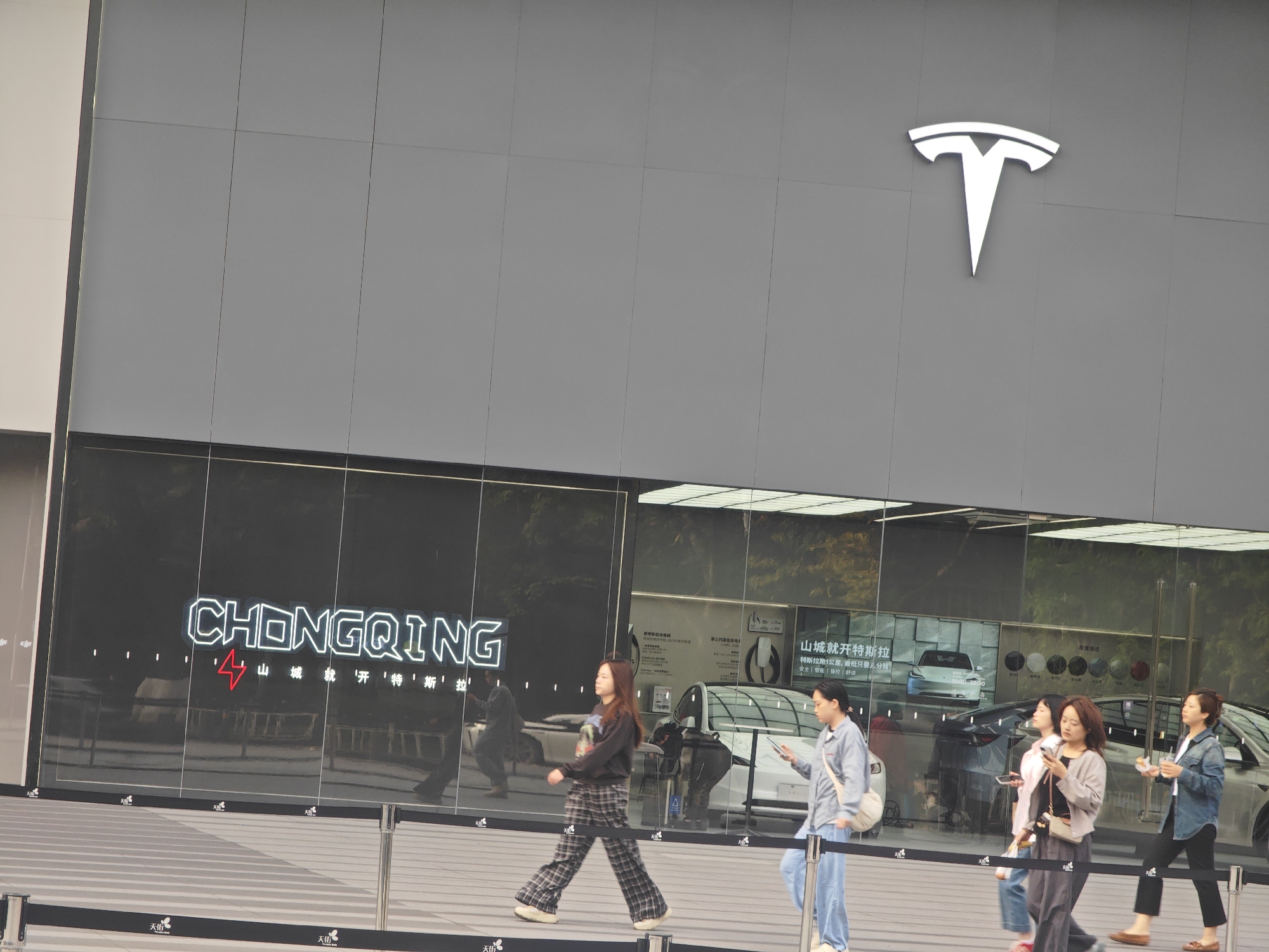 The image displays the exterior of a modern Tesla showroom in Chongqing, China, during daylight hours. The building features a minimalist design with a large, dark gray upper facade, prominently adorned with a white, illuminated Tesla logo. The ground floor is comprised of expansive glass windows, which reflect the surroundings and offer glimpses into the showroom's interior.
On the glass windows, large, neon-style text spells out "CHONGQING," accompanied by Chinese characters that read "山城就开特斯拉" (Mountain City opens Tesla), indicating the specific location. Inside the showroom, through the reflections, a silver Tesla vehicle is partially visible, alongside what appear to be charging stations labeled "第三代电" (Third generation electric). Promotional posters also display information such as "特斯拉跑1公里,最低只要儿分钱" (Tesla runs 1 kilometer, minimum only a few fen) and keywords like "安全|智能|操控|舒适" (Safety | Intelligence | Handling | Comfort).
Along the sidewalk in front of the building, a black barrier features the repeated text "天街" (Tian Jie). Several pedestrians, predominantly women, are seen walking casually from left to right on the sidewalk. They are dressed in everyday clothing, with some individuals looking at their mobile phones. Their expressions appear neutral, suggesting they are either passing by or engaging in window shopping in this urban commercial setting. The soft natural light indicates it is daytime, possibly an overcast day or late afternoon.