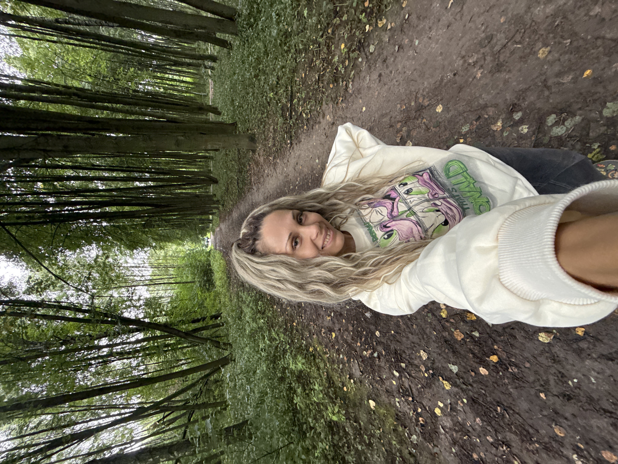 A woman with long, wavy blonde hair smiles at the camera in this selfie, taken from a low angle. She is lying down on a dirt path in a forest, with her head propped up. She wears a white hooded sweatshirt with a graphic print on the front, depicting cartoon aliens and text that appears to read "OMWAD." Her eyes are open, and she has a joyful expression.
The setting is a dense forest with tall trees lining a narrow dirt path. The trees are closely packed, creating a tunnel-like effect. Sunlight filters through the green canopy, casting dappled light on the path. Fallen leaves are scattered across the ground, suggesting it may be autumn. The path ahead is visible, leading deeper into the woods. The overall atmosphere is peaceful and natural. There are no obvious cues for the time of day or specific weather conditions, but the lighting suggests it is daytime. The location is identified as Rusanovo, Russia.
