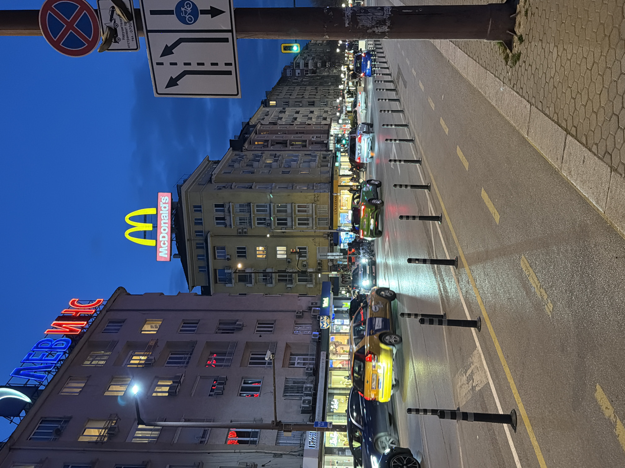 The image depicts an urban street scene at dusk or early night in Sofia, Bulgaria, characterized by a deep blue sky and illuminated artificial lights.
The primary location identifier is the large, illuminated neon sign reading "ЛЕВ ИНС" (LEV INS), a Bulgarian insurance company, atop a multi-story building on the left. Adjacent to it, the prominent yellow McDonald's "M" logo and "McDonald's" text sign are visible on a slightly smaller building further down the street. The street itself is wet, reflecting the numerous vehicle lights and overhead street lamps, suggesting recent rain or damp conditions.
Multiple vehicles are in motion on the road, with some exhibiting motion blur. A yellow and blue taxi is prominent in the middle ground, moving away from the viewer. Other vehicles include dark sedans and SUVs. The road features clearly marked lanes with white and yellow lines. A row of black and grey bollards separates the main traffic lanes from what appears to be a designated bicycle lane or shoulder on the right side of the road.
On the right, a utility pole is visible, bearing multiple road signs. These include a "No Stopping" sign, a tow-away zone sign with a graphic of a tow truck and Bulgarian text indicating penalties for violators (Нарушители отстраняват пр тел. 0700...), and a directional lane sign indicating one lane for left turn/straight, one for straight, and one for straight with a bicycle lane designation. A green traffic light is visible further down the street on the right.
The surrounding environment consists of multi-story residential and commercial buildings lining both sides of the street, many with illuminated windows and ground-floor storefronts, including an L'Occitane store. The sidewalk on the right features hexagonal patterned paving stones. No individual pedestrians are clearly discernible, but the presence of shops suggests an active urban area.