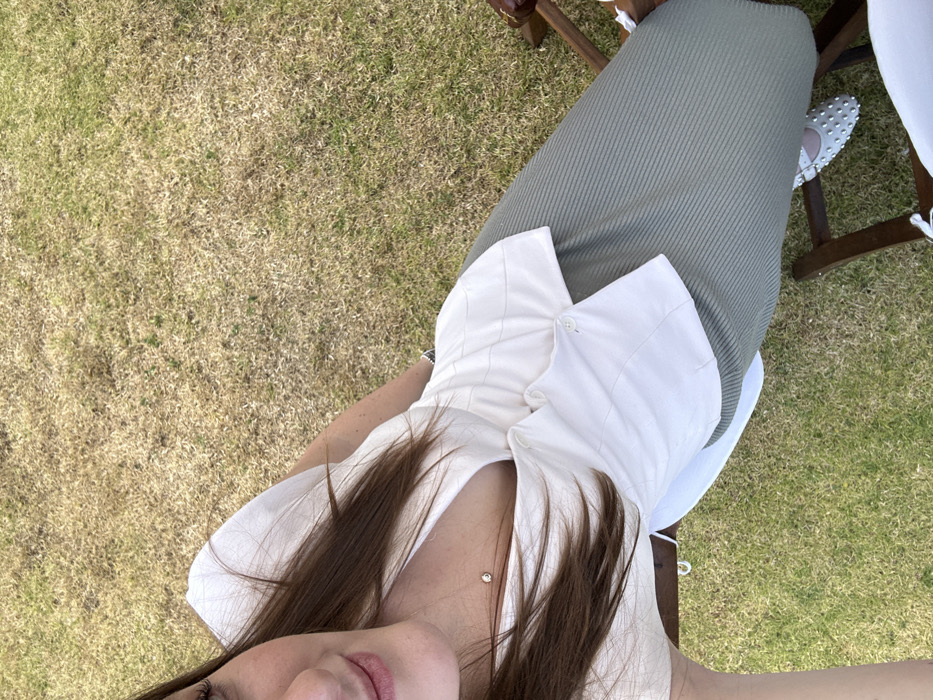 A high-angle photograph captures a female individual seated outdoors in the city of Unknown, Unknown. The subject's chest, abdomen, legs, and feet are visible, along with a partial view of her chin, lips, nose, and lower right eye in the upper left corner. She has long brown hair. She is wearing a white, collared V-neck top with a quilted stitch pattern and a single button closure at the waist, an olive green vertically ribbed midi skirt, and white ballet flats adorned with silver studs. A silver chain necklace with a small, clear stone pendant is visible on her chest, and a metallic bracelet or watch is on her left wrist. She is positioned between two partially visible wooden chairs, one on each side, both featuring white fabric ties. The environment consists of an outdoor lawn with a mix of green and predominantly dry, yellowish-brown grass. The individual is stationary, appearing to take a selfie. No other people or interactions are present.