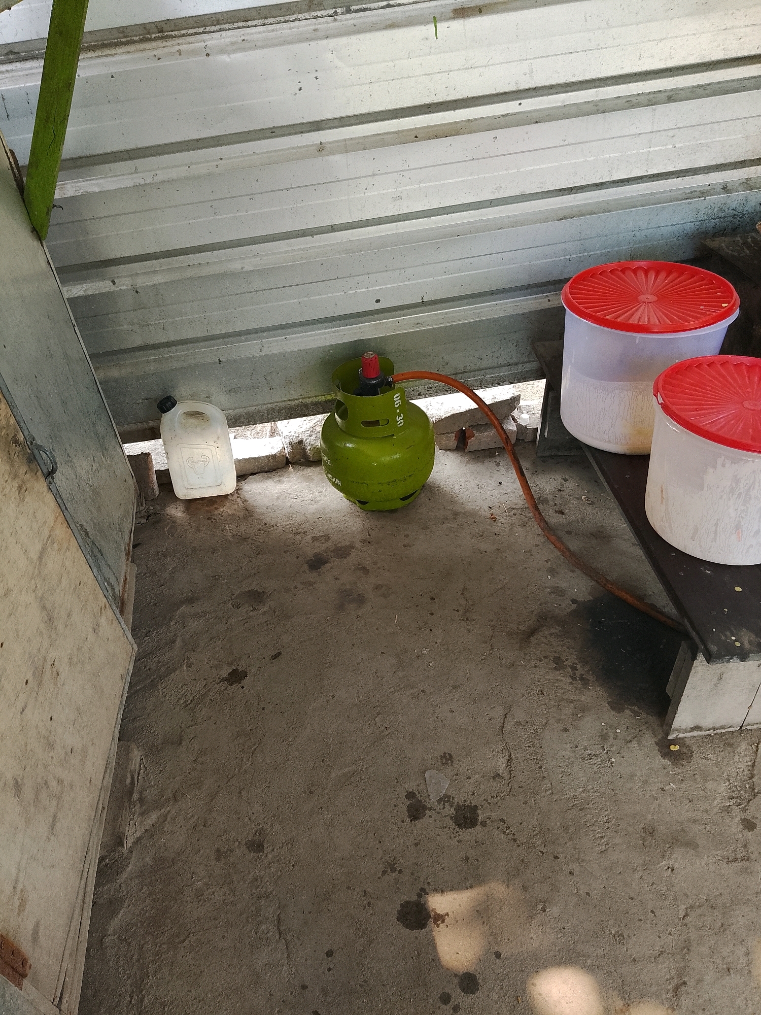 In Banaran, Indonesia, a green gas cylinder with a red regulator is positioned on a concrete floor. An orange hose connects the regulator to an unseen appliance. To the left of the gas cylinder, a white plastic jerrycan with a black cap rests on the ground. In the background, two large translucent plastic containers with red lids are stacked on a raised platform. The scene appears to be an outdoor or semi-enclosed space, possibly a storage area, with a corrugated metal roof overhead and a wooden panel to the left. The ground is concrete with some dark stains.
