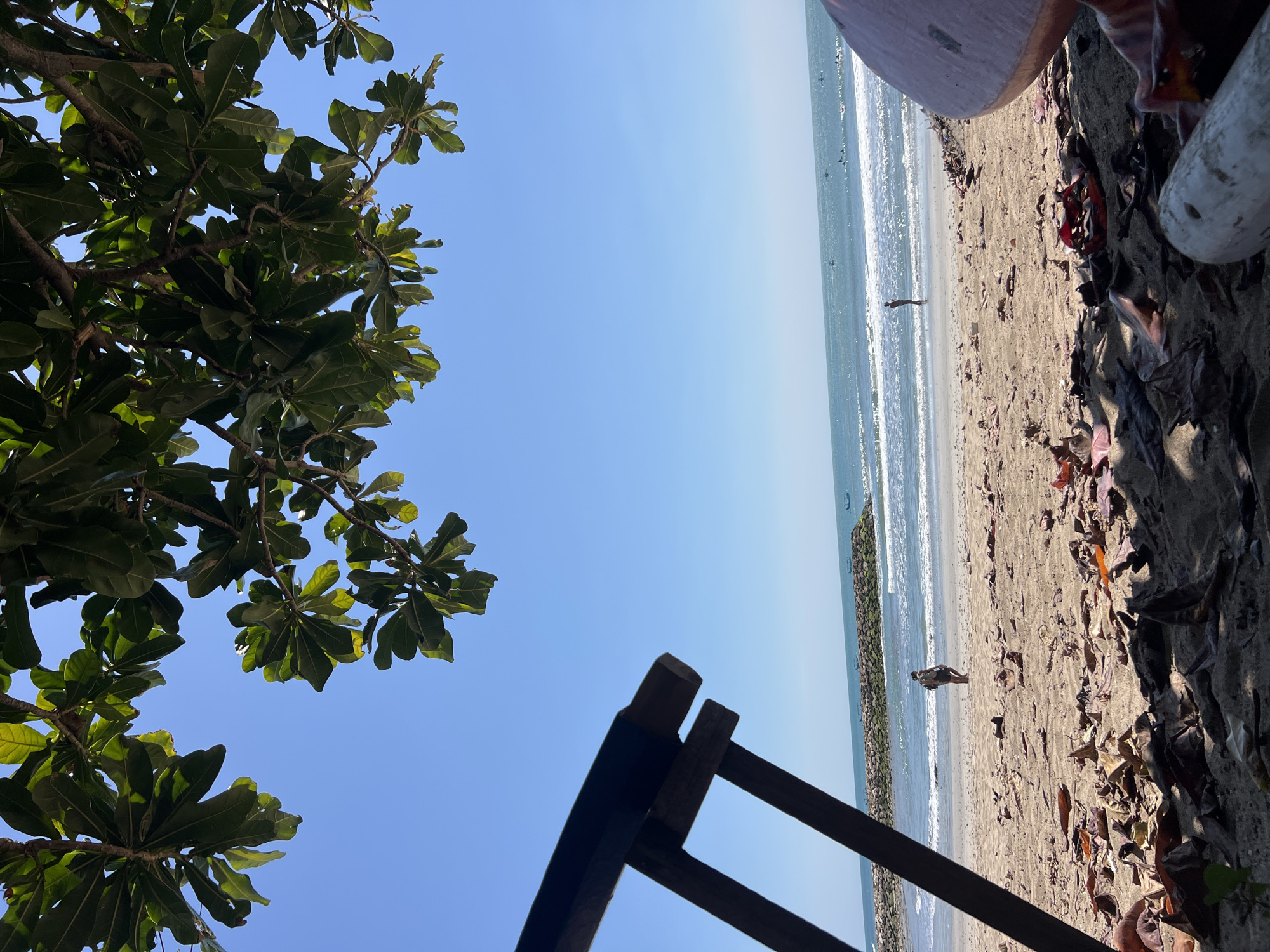 An eye-level shot taken on a sunny day shows a beach in Kuta, Indonesia. In the foreground, a wooden chair is partially visible on the left, and a pink, rounded object on the right, both casting shadows on the sandy ground littered with dried leaves. A person with a backpack is walking on the beach near the water, which has small waves. A stone jetty stretches out into the ocean. Further out, several small boats are visible on the sea, and a person is standing near the water's edge. Lush green leaves from a tree hang from the top of the frame against a clear blue sky.
