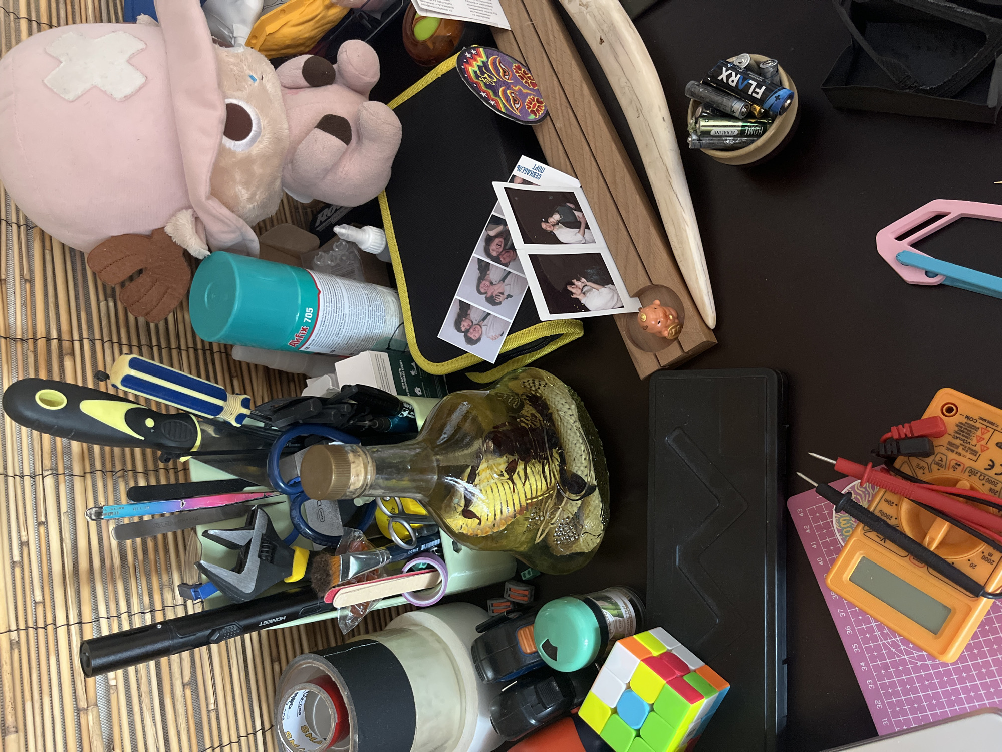 The image shows a cluttered desk or workspace. The background consists of bamboo blinds. On the desk, there are various objects including a Rubik's Cube, a bottle containing a preserved snake, several tools such as an adjustable wrench and screwdrivers, a stuffed animal, a can of spray, a multimeter, and a collection of batteries in a small bowl. There are also several printed photographs displayed on a wooden holder. The overall impression is one of a personal workspace where various hobbies or projects might be undertaken. The image does not provide any information about the location in Moscow, Russia.
