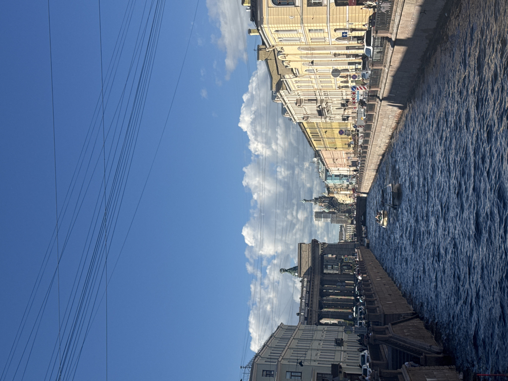 This media file captures a scenic view of a canal in Saint Petersburg, Russia. The scene is dominated by the calm, blue waters of the canal on the right side, reflecting the sky and surrounding architecture. Along the left bank of the canal, a row of ornate, multi-story buildings with yellow and pastel facades line the street. These buildings feature classical architectural elements, including balconies, arched windows, and decorative detailing. A pedestrian walkway runs along the canal, bordered by a dark railing, with various signs and traffic lights visible, suggesting an urban environment. Cars are parked along the street, and a few people are visible strolling on the walkway.
In the distance, more buildings and architectural landmarks are visible, hinting at the city's rich history and grandeur. A bright blue sky with scattered white clouds stretches across the top half of the image. A prominent feature on the left side of the image is a set of numerous dark power lines stretching across the sky, adding a modern, man-made element to the otherwise historical cityscape. The lighting suggests it is daytime, likely a sunny day with some cloud cover. The overall mood is serene and picturesque, showcasing the beauty of Saint Petersburg's urban canals and architecture.