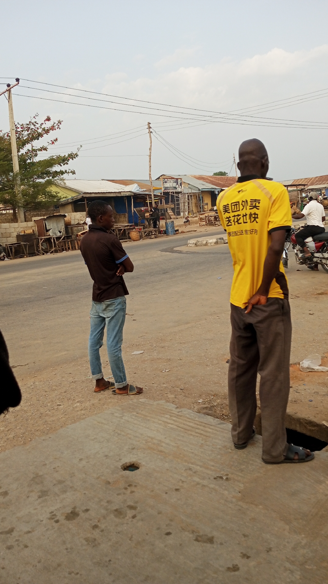 The image captures a street scene in Zango-ndaji, Nigeria. Two men are the primary subjects. One man wears a brown, collared shirt and light blue jeans, standing with his arms crossed. The other man is wearing a bright yellow t-shirt with Chinese characters, coupled with grey trousers. He is standing with his hands behind his back. A motorcycle with a rider in a white shirt is visible in the background. The environment includes buildings with corrugated metal roofs lining the street, power lines overhead, and a concrete structure with a hole near the foreground.