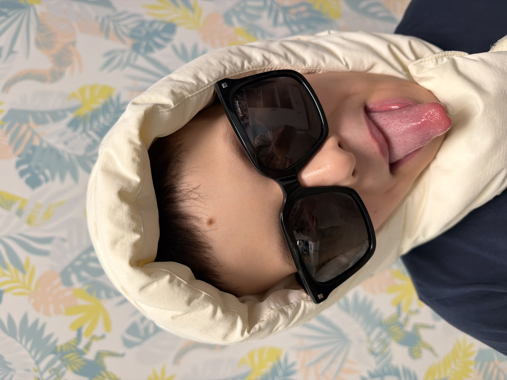 A person wearing black sunglasses and a light-colored hooded jacket is lying on a bed with a floral pattern sheet. The person is sticking their tongue out. The photograph was not taken in Votkinsk, Russia.