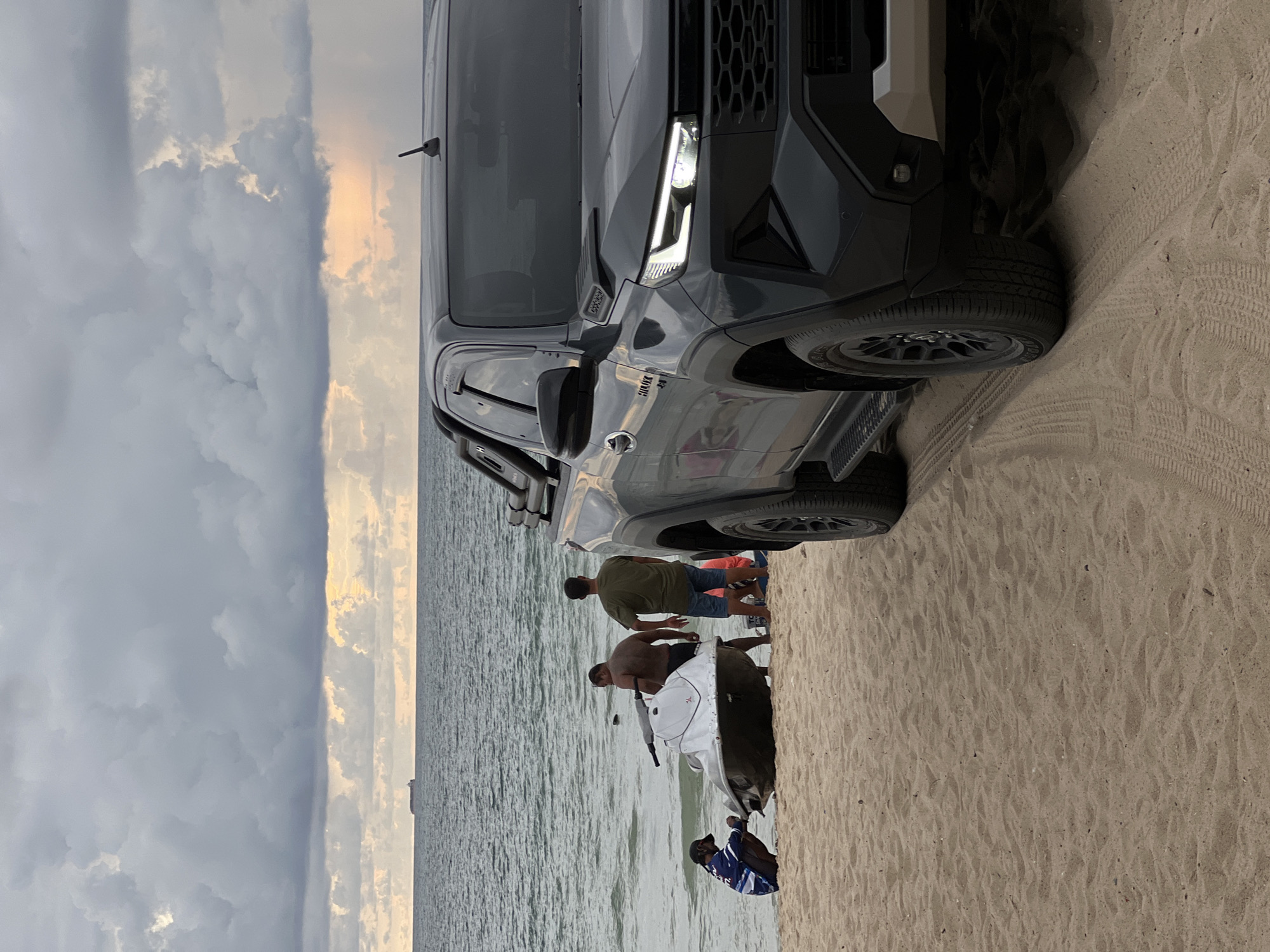 This is an eye-level, medium shot of a dark gray Toyota Hilux pickup truck parked on a sandy beach. The truck is angled towards the viewer and occupies the right side of the frame. In the background, the ocean stretches to the horizon under a dramatic, cloudy sky with hints of sunlight breaking through. A large cargo ship is visible on the distant horizon. On the beach, to the left of the truck, three men are interacting with a white jet ski. One man is kneeling next to the jet ski, appearing to adjust or inspect it. Another man stands behind the jet ski, and a third man stands to his right, facing away from the camera. The environment is a beach with sand in the foreground and water in the background. The sky is overcast with some patches of light, suggesting the possibility of rain.