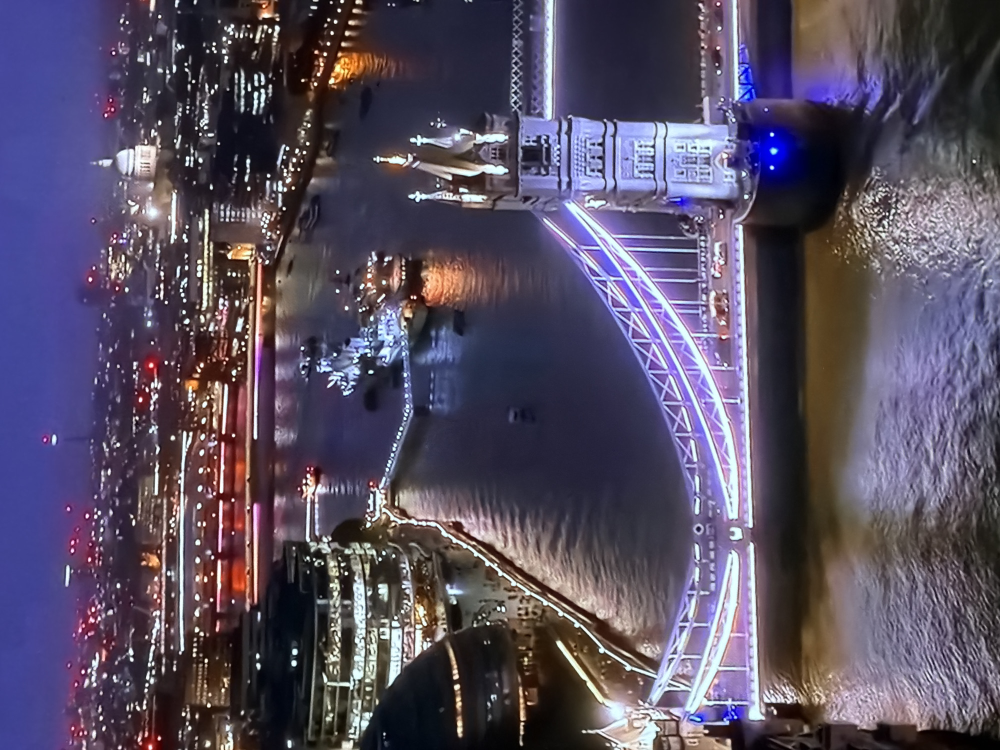An aerial night-time view captures London, United Kingdom, showcasing iconic landmarks. The central feature is Tower Bridge, spanning the River Thames and illuminated in vibrant purple and white lights. Vehicular traffic is visible on the bridge, and several boats navigate the water below, including the historic warship HMS Belfast docked upstream. On the river's south bank, the distinct, rounded glass building of City Hall is visible. In the distant background, the prominent dome of St Paul's Cathedral rises above the illuminated cityscape, which extends across the horizon with numerous lights. The sky is dark, enhancing the brilliance of the artificial lighting reflecting on the water's surface. The location in the city of Maidstone, United Kingdom, is also noted.